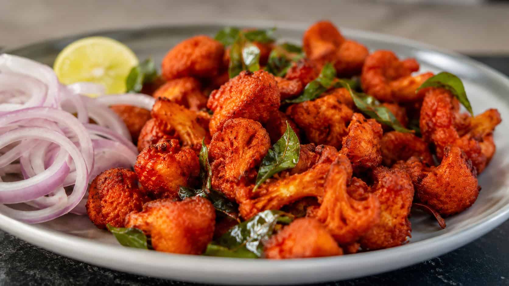 spicy red gobhi 65 crispy cauliflower with curry leaves in a wide horizontal shot, served on a plate with lemon and onion rings, warm natural light, shallow depth of field, landscape orientation