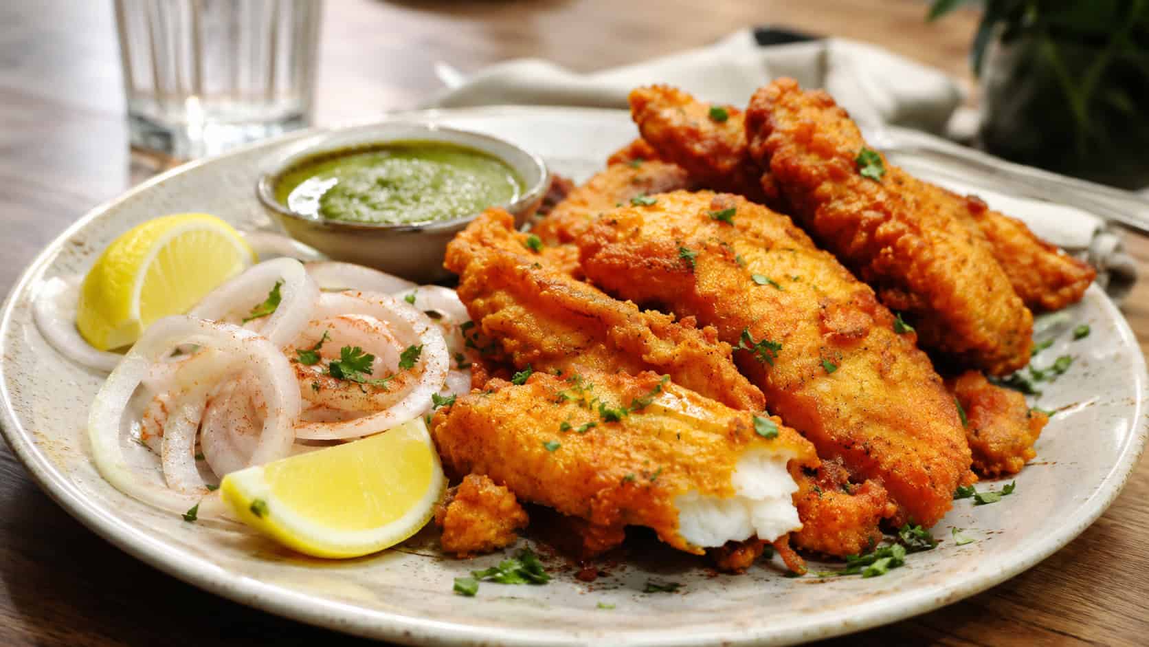 golden crispy fish amritsari fritters in a wide horizontal shot, served on a plate with onion rings, lemon wedges, and green chutney, warm natural light, shallow depth of field, landscape orientation