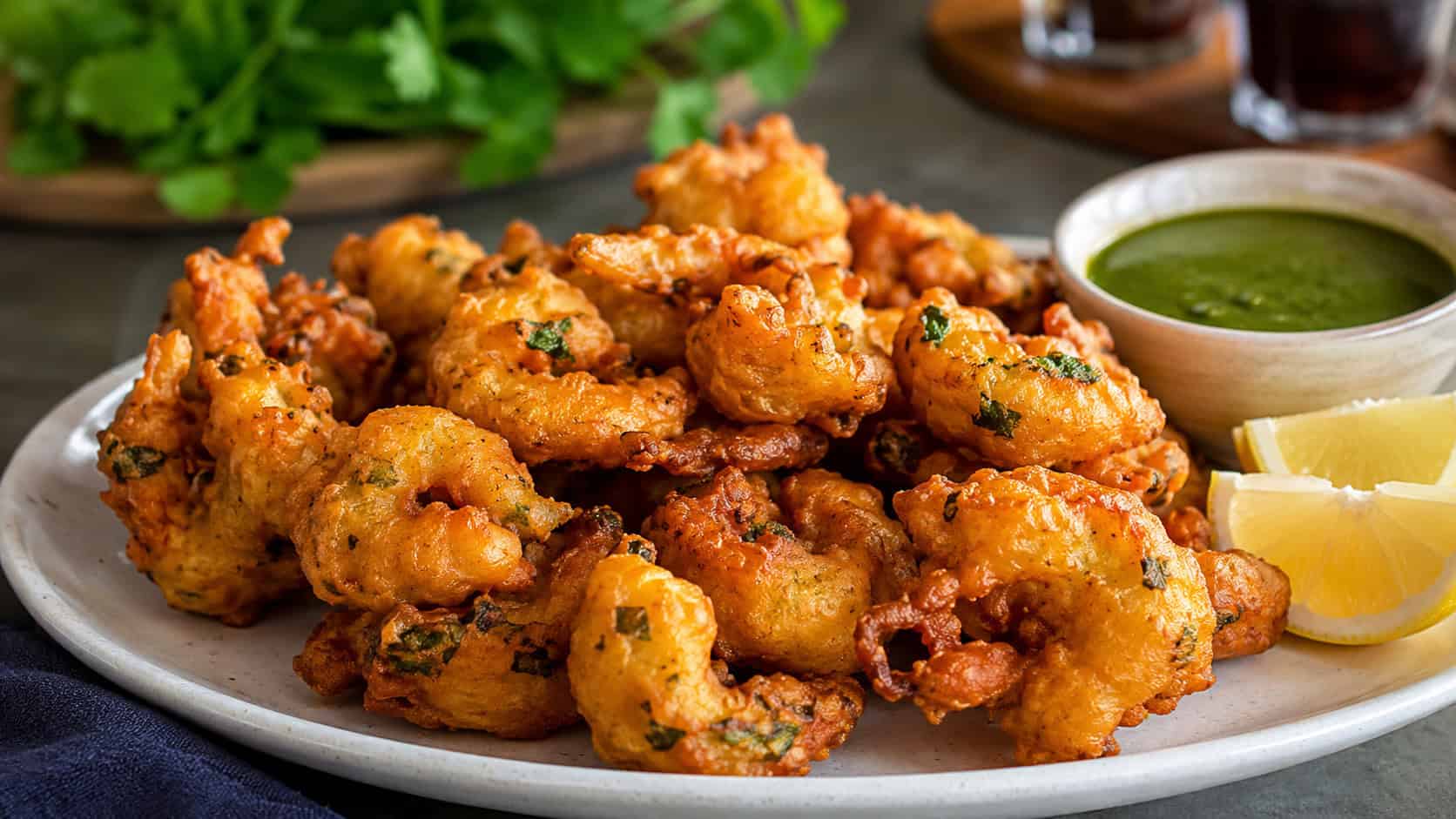 crispy golden shrimp pakora fritters in a wide horizontal shot, piled on a plate with mint chutney and lemon wedges, warm natural light, shallow depth of field, landscape orientation