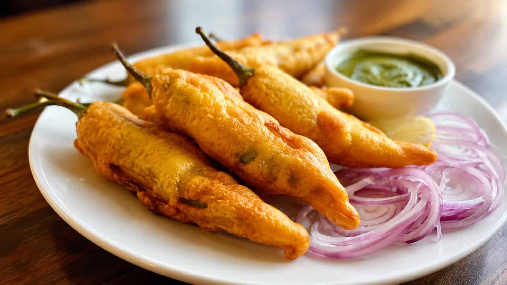 golden mirchi bajji battered fried chilies in a wide horizontal shot, served on a plate with onion slices and chutney, warm natural light, shallow depth of field, landscape orientation
