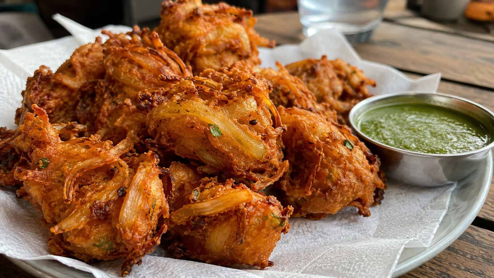 crispy golden onion bhaji fritters in a wide horizontal shot, piled on a plate lined with paper and served with green chutney, warm natural light, shallow depth of field, landscape orientation