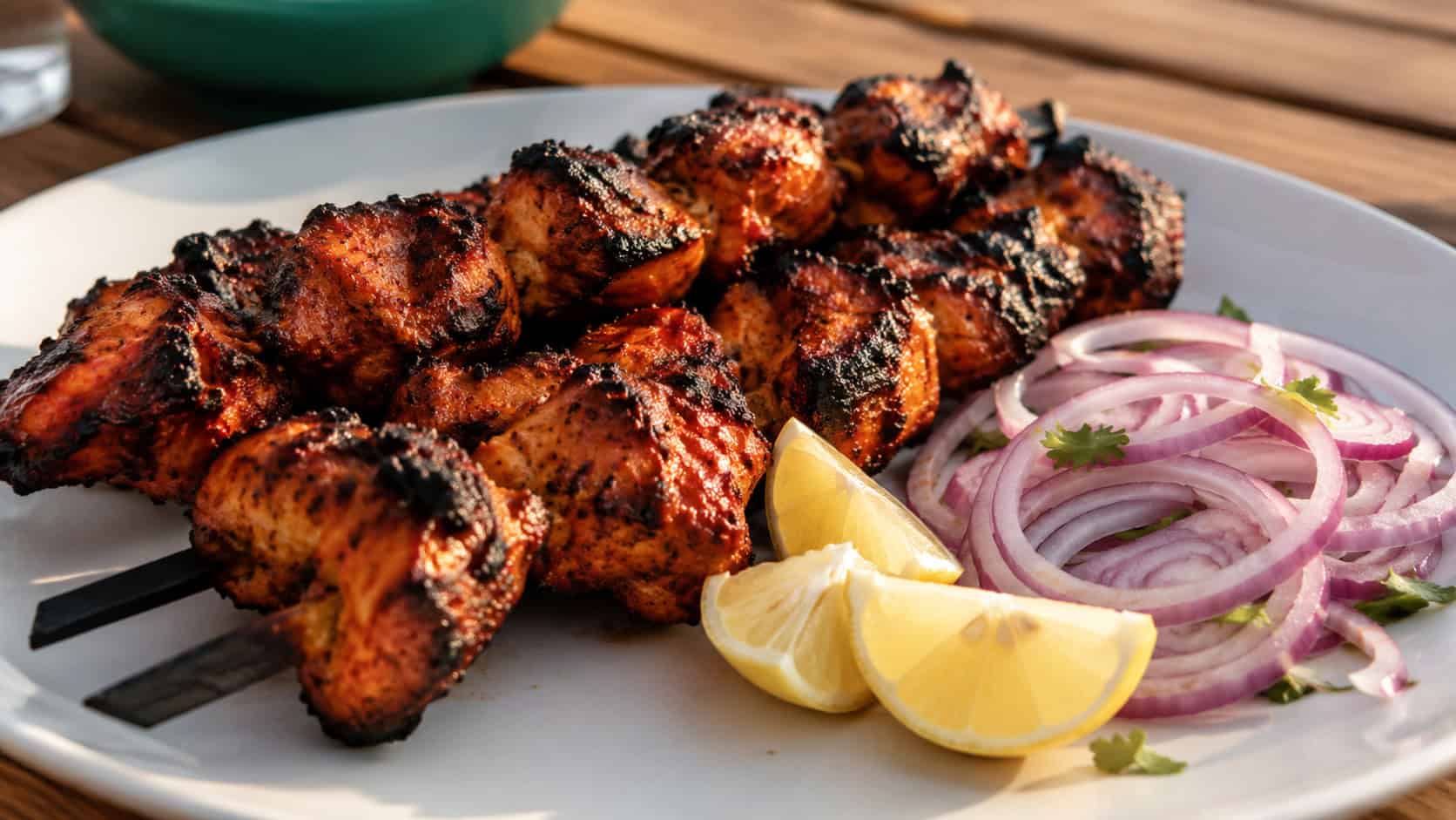 charred chicken tikka pieces with smoky edges on skewers in a wide horizontal shot, plated with red onion rings and lemon wedges, warm natural light, shallow depth of field, landscape orientation
