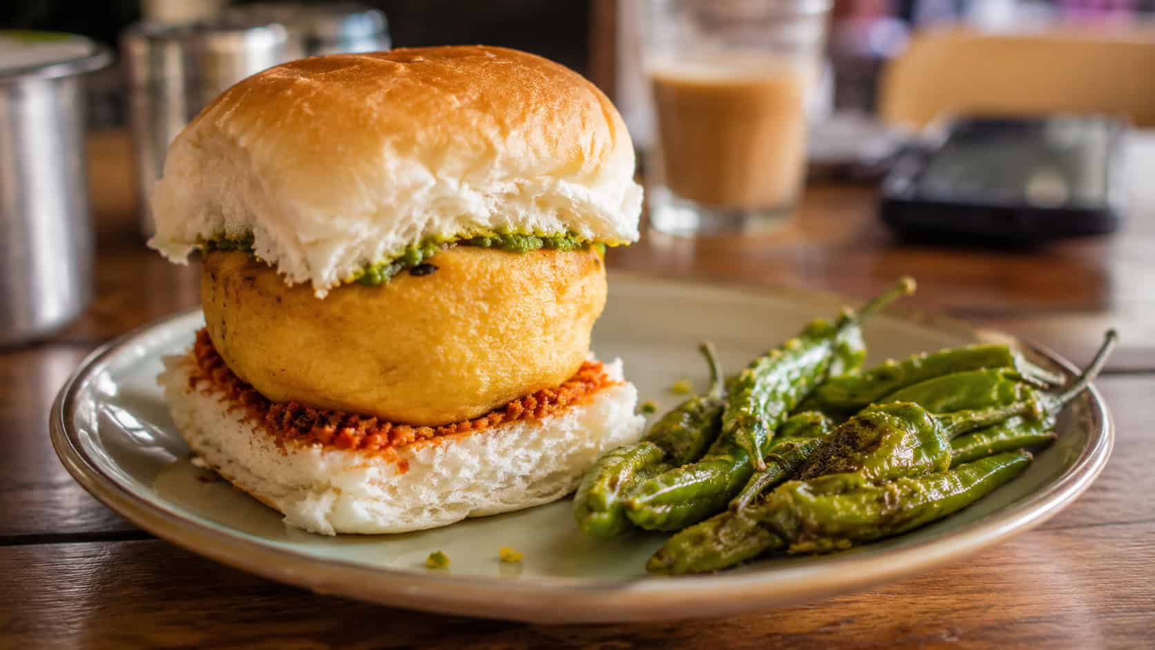 vada pav potato slider in a soft bun with chutney in a wide horizontal shot, served on a plate with fried green chilies on a casual table, warm natural light, shallow depth of field, landscape orientation