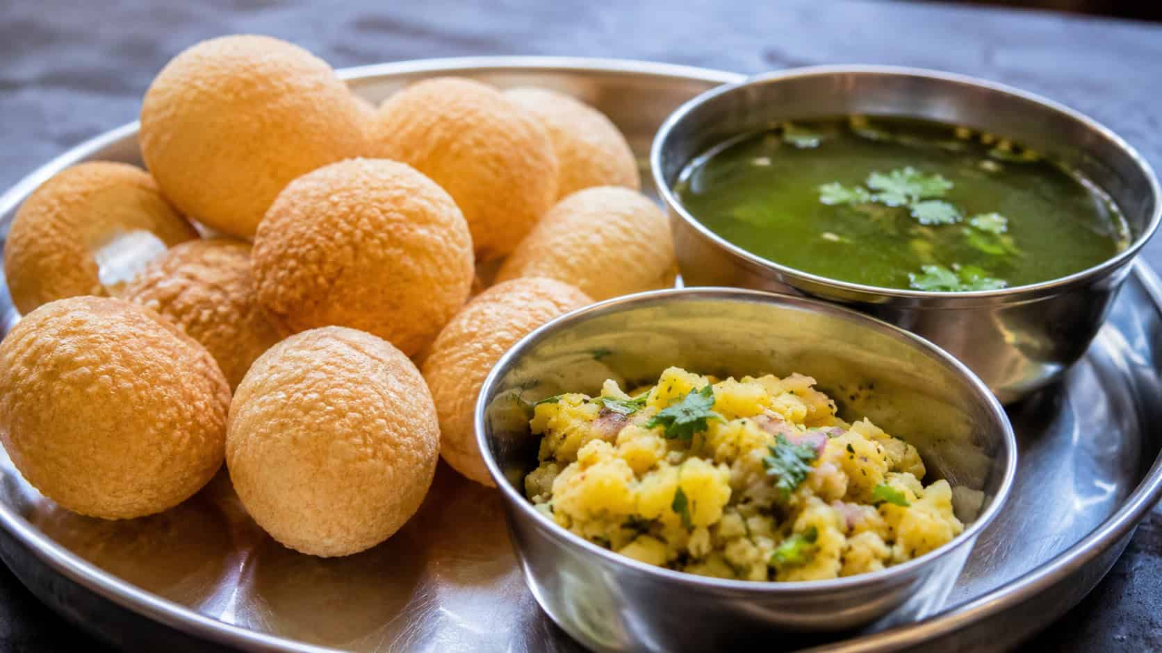 pani puri crisp shells alongside a bowl of green spiced water and potato filling in a wide horizontal shot, arranged on a metal thali platter, bright natural light, shallow depth of field, landscape orientation