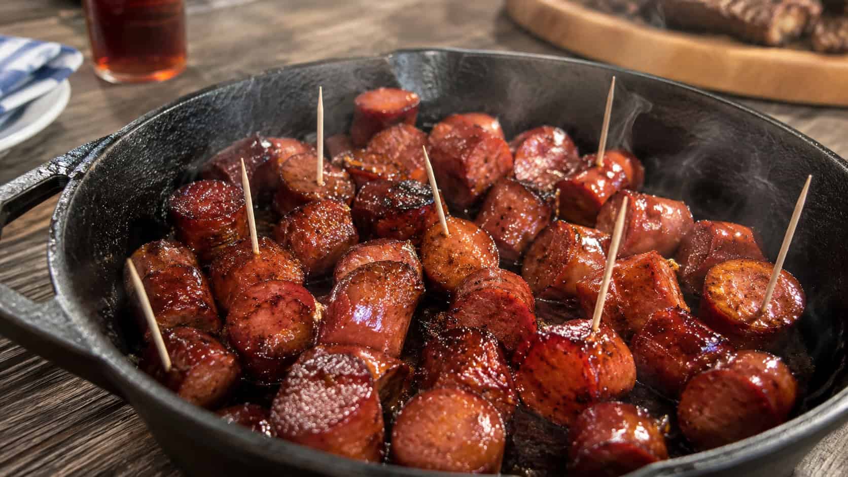 a wide skillet of glossy maple-glazed smoked sausage pieces with a sticky sheen, toothpicks tucked in, faint smoke, on a rustic table, warm natural light, shallow depth of field, landscape orientation