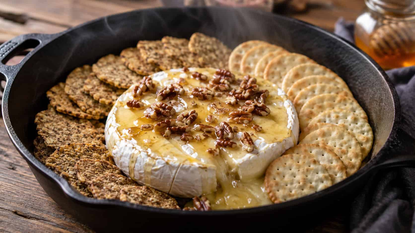 a wide cast iron skillet with a smoked gooey wheel of brie drizzled with honey and pecans, surrounded by crackers, faint smoke, on a rustic wooden table, warm natural light, shallow depth of field, landscape orientation