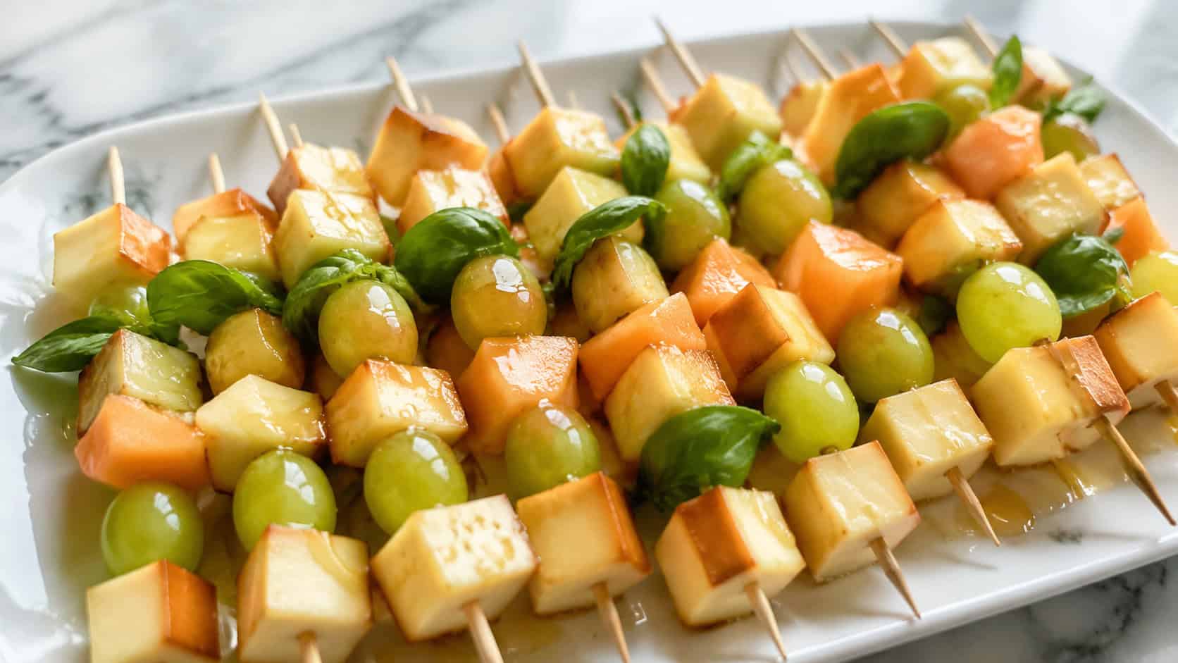 a wide platter of skewers alternating smoked cheese cubes, green grapes, and orange melon with basil leaves, drizzled with honey, on a marble surface, bright natural light, shallow depth of field, landscape orientation