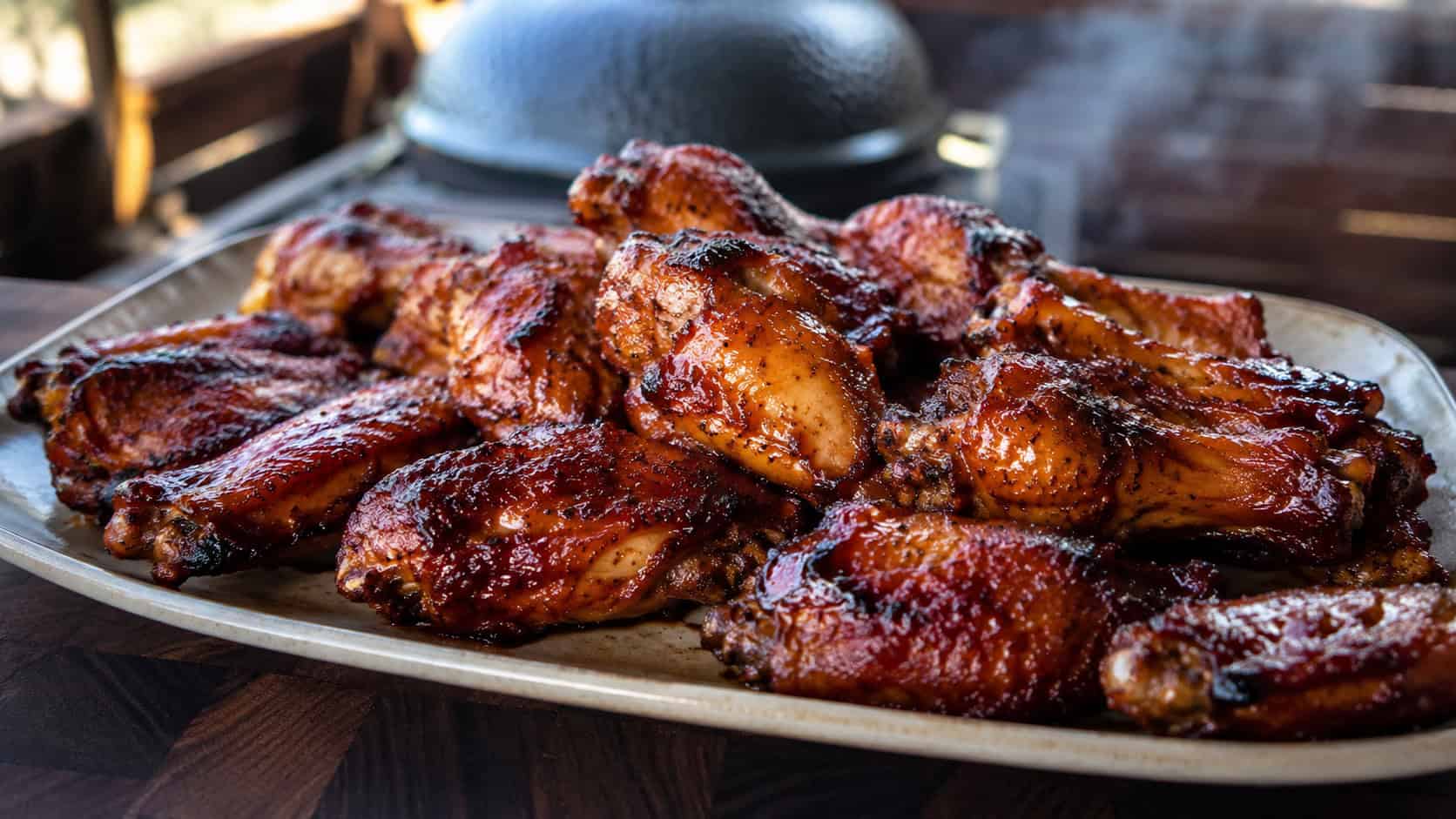 a wide platter of glossy smoked chicken wings glazed with BBQ sauce and a crispy skin, faint smoke in the background, on a dark wooden board, warm natural light, shallow depth of field, landscape orientation