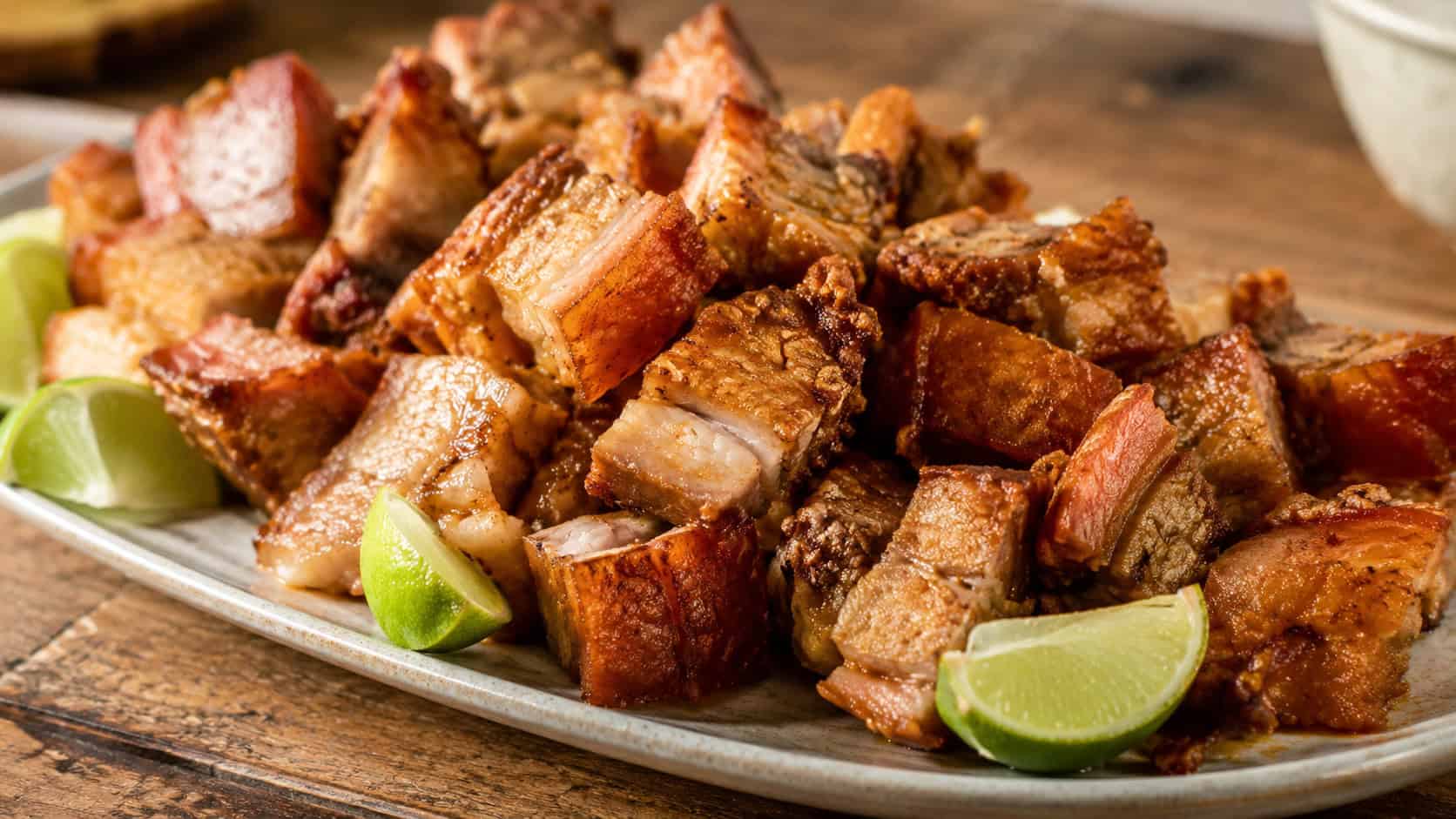 a wide platter of crispy golden fried pork chunks with crackling edges, lime wedges tucked around them, on a rustic wooden board, warm natural light, shallow depth of field, landscape orientation