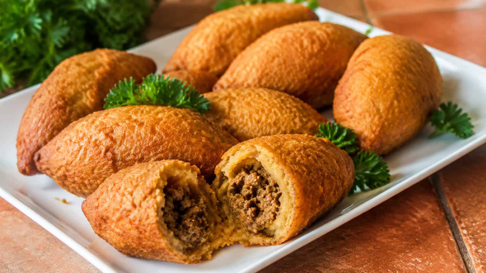several golden brown fried alcapurria fritters arranged on a wide white platter, one split open to reveal seasoned ground beef filling, on a warm terracotta surface, garnished with parsley, natural light, shallow depth of field, landscape orientation
