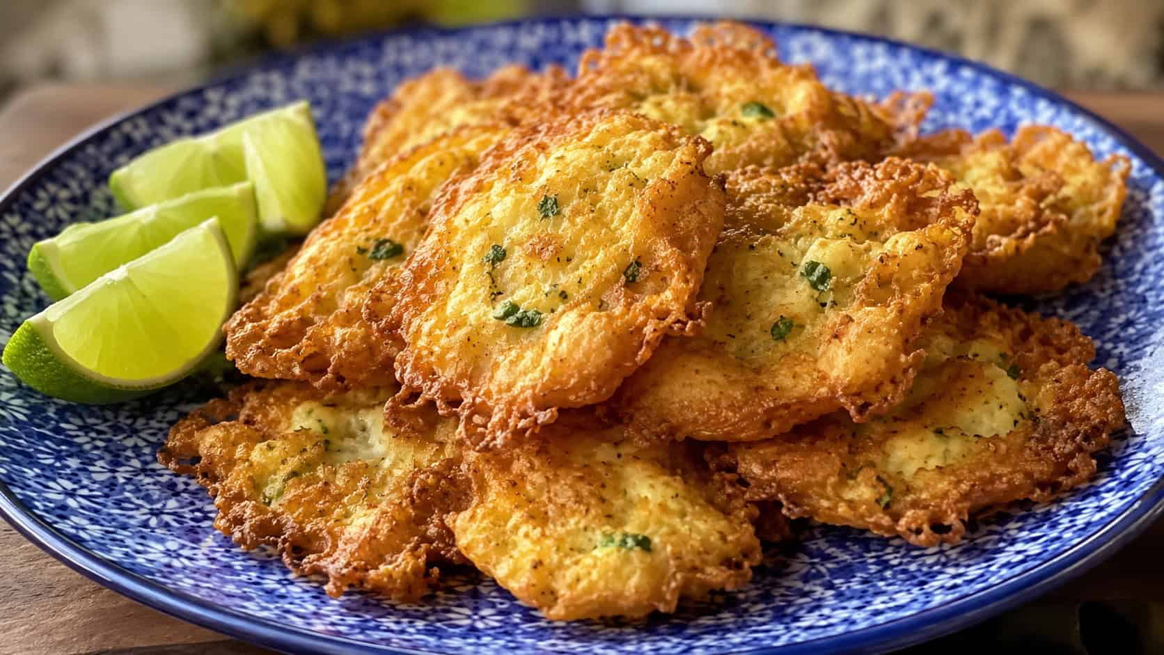 a wide plate of thin crispy golden codfish fritters with lacy edges, stacked loosely, on a blue patterned ceramic plate, lime wedges to the side, bright natural light, shallow depth of field, landscape orientation