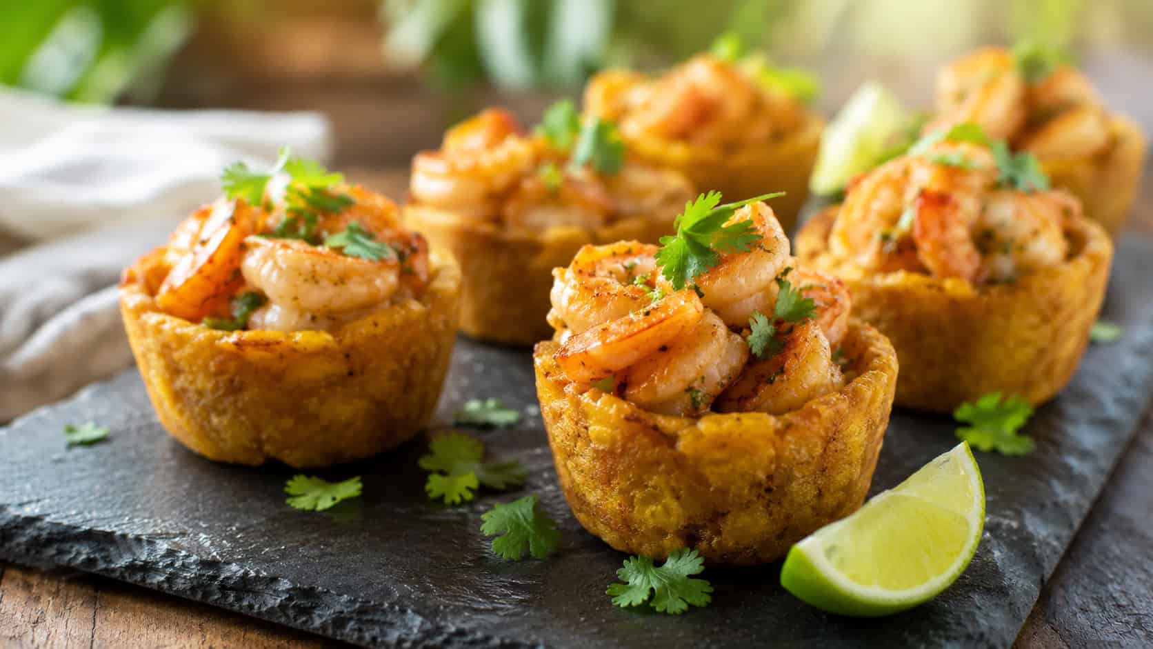several small mashed plantain mofongo cups filled with garlicky shrimp, arranged on a wide slate platter, garnished with cilantro and lime, warm natural light, shallow depth of field, landscape orientation