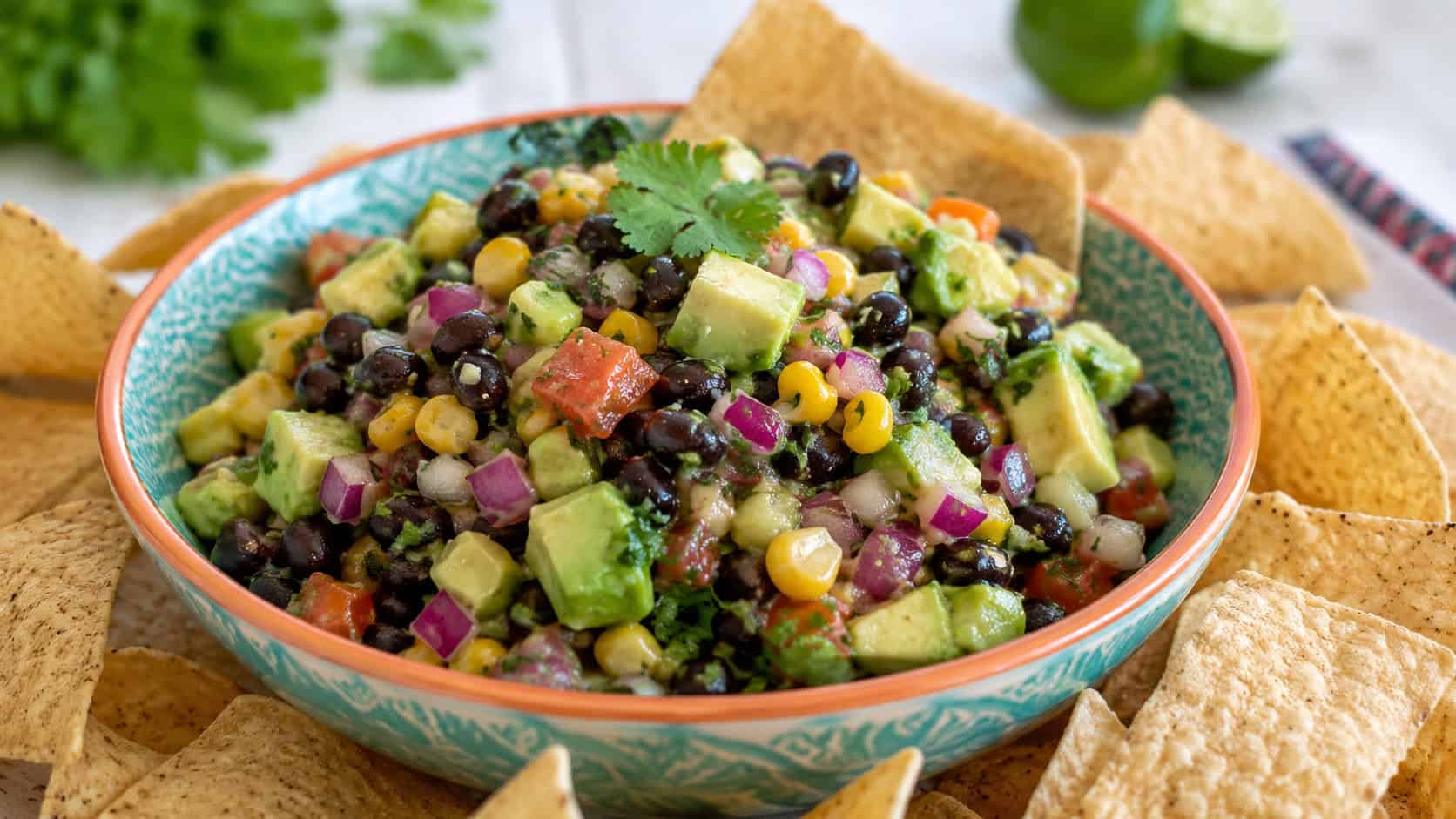 a colorful bowl of avocado black bean salsa with corn, red onion, and cilantro, surrounded by tortilla chips, in a wide horizontal shot on a bright table, natural light, shallow depth of field, landscape orientation