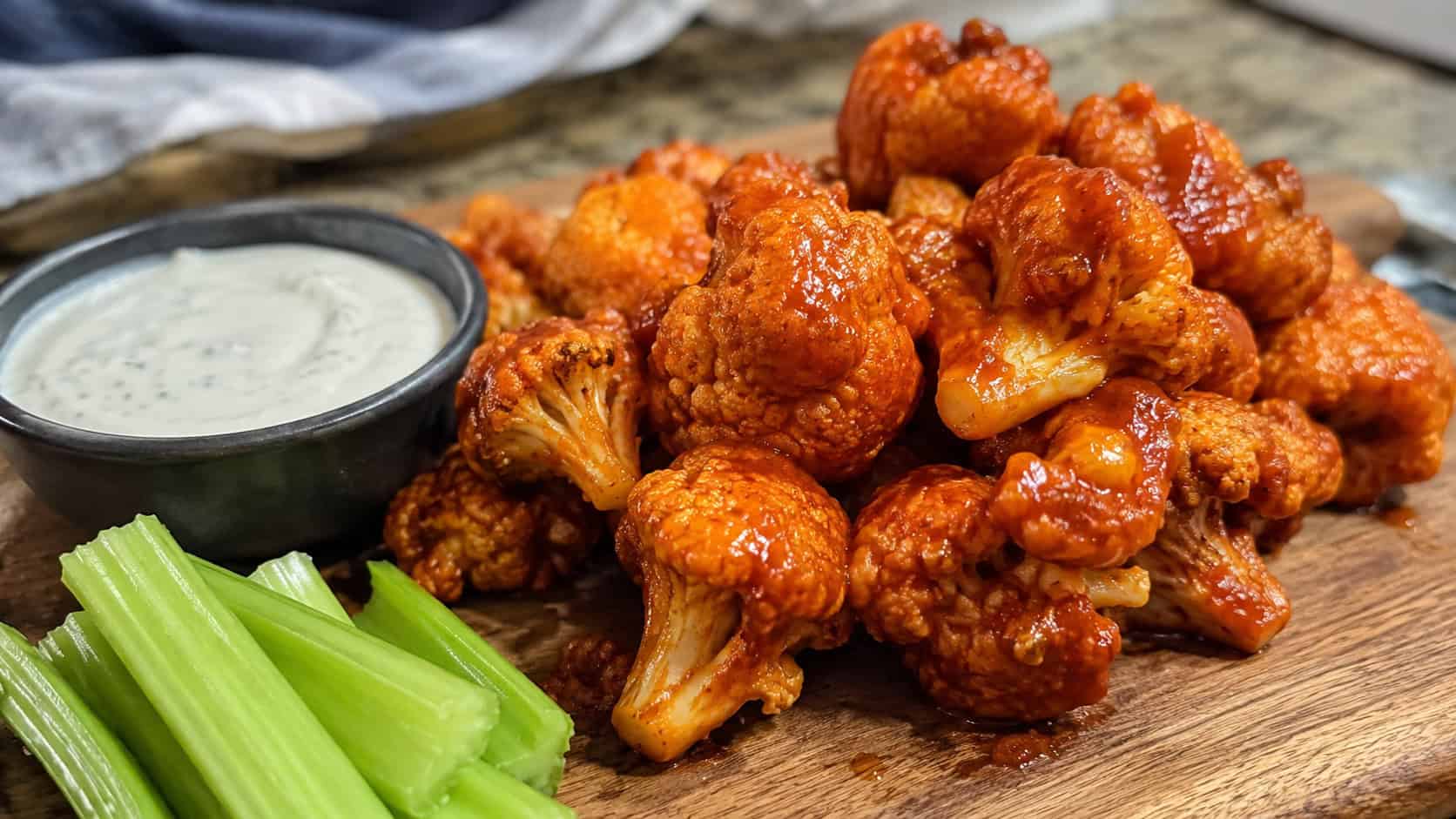 a pile of crispy buffalo cauliflower wings glazed in orange sauce with celery sticks and ranch on the side, in a wide horizontal shot on a board, natural light, shallow depth of field, landscape orientation