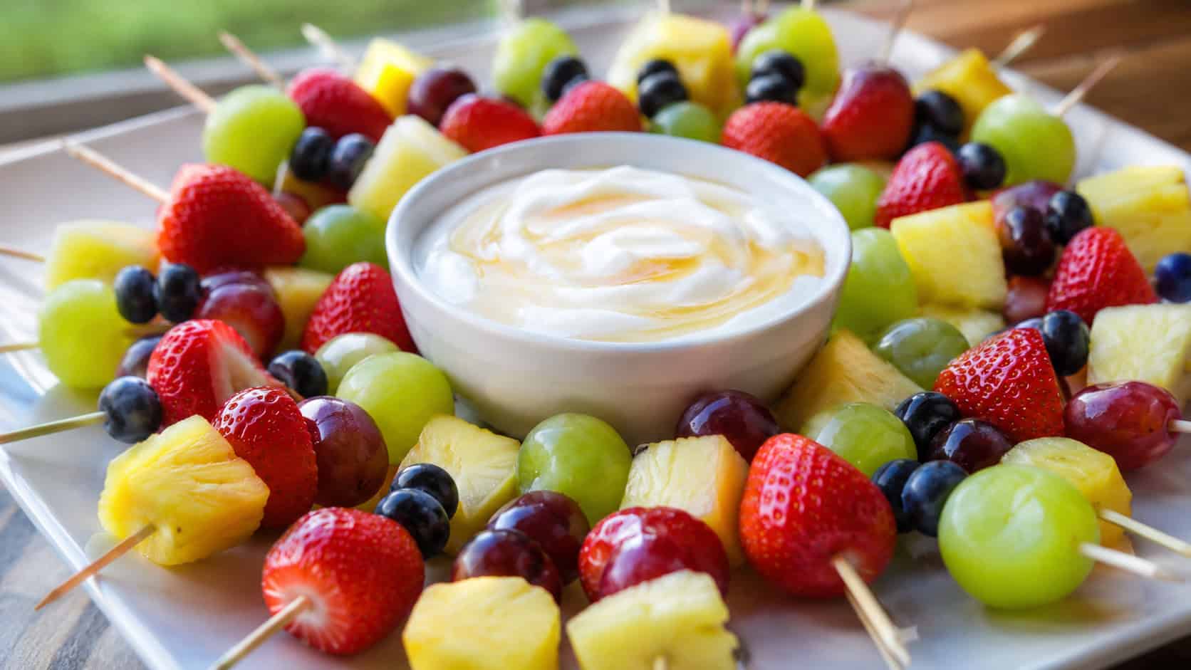 colorful mini fruit skewers with strawberries, pineapple, grapes, and blueberries around a bowl of honey yogurt dip, in a wide horizontal shot on a bright platter, natural light, shallow depth of field, landscape orientation