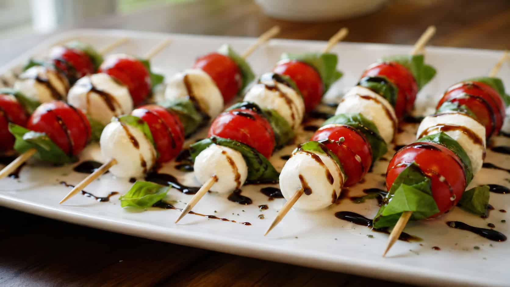 caprese skewers with cherry tomatoes, mozzarella, and basil drizzled with balsamic glaze lined up on a white platter, in a wide horizontal shot, natural light, shallow depth of field, landscape orientation
