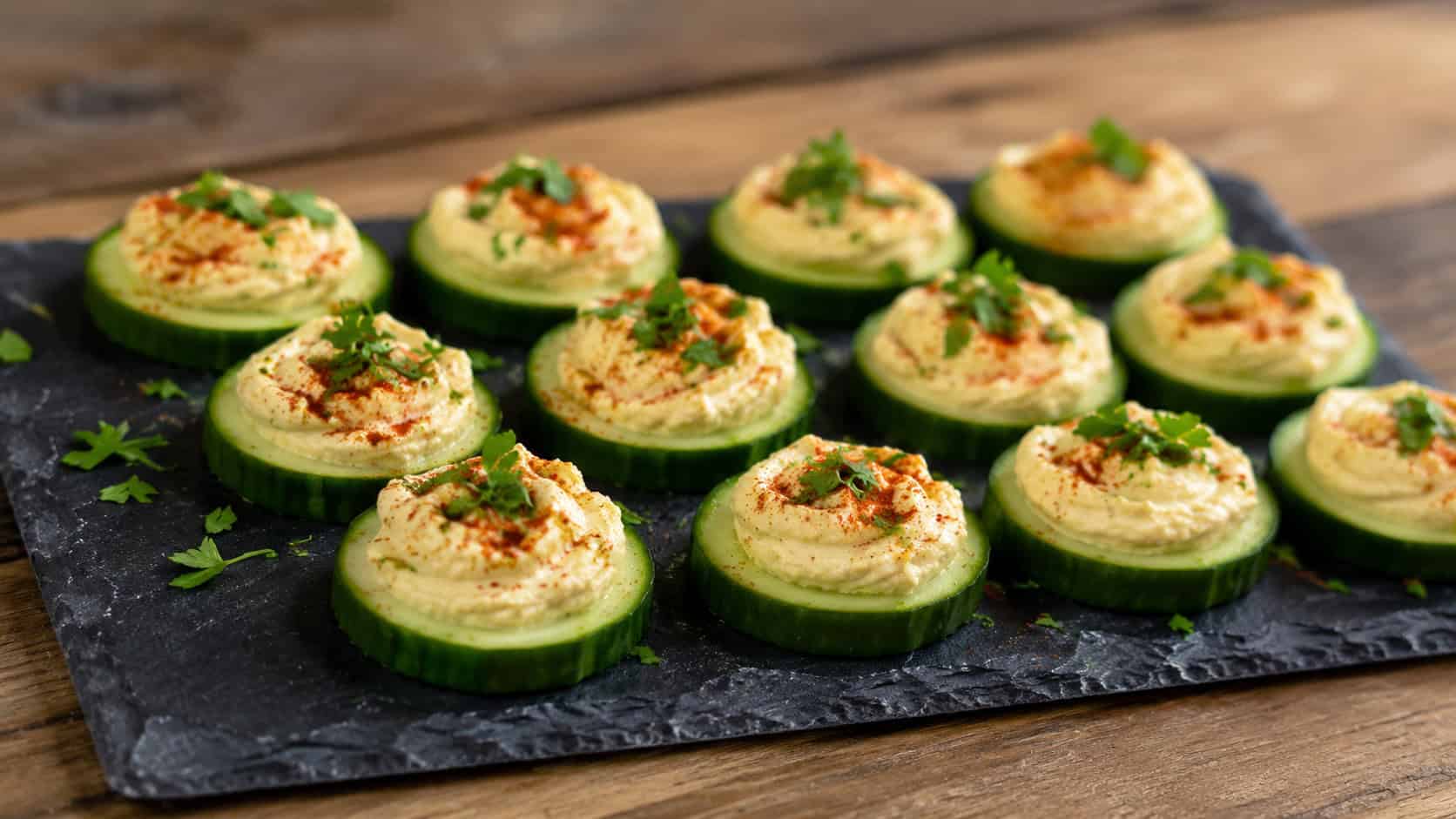 cucumber rounds topped with creamy hummus, paprika, and parsley arranged on a slate board, in a wide horizontal shot, natural light, shallow depth of field, landscape orientation