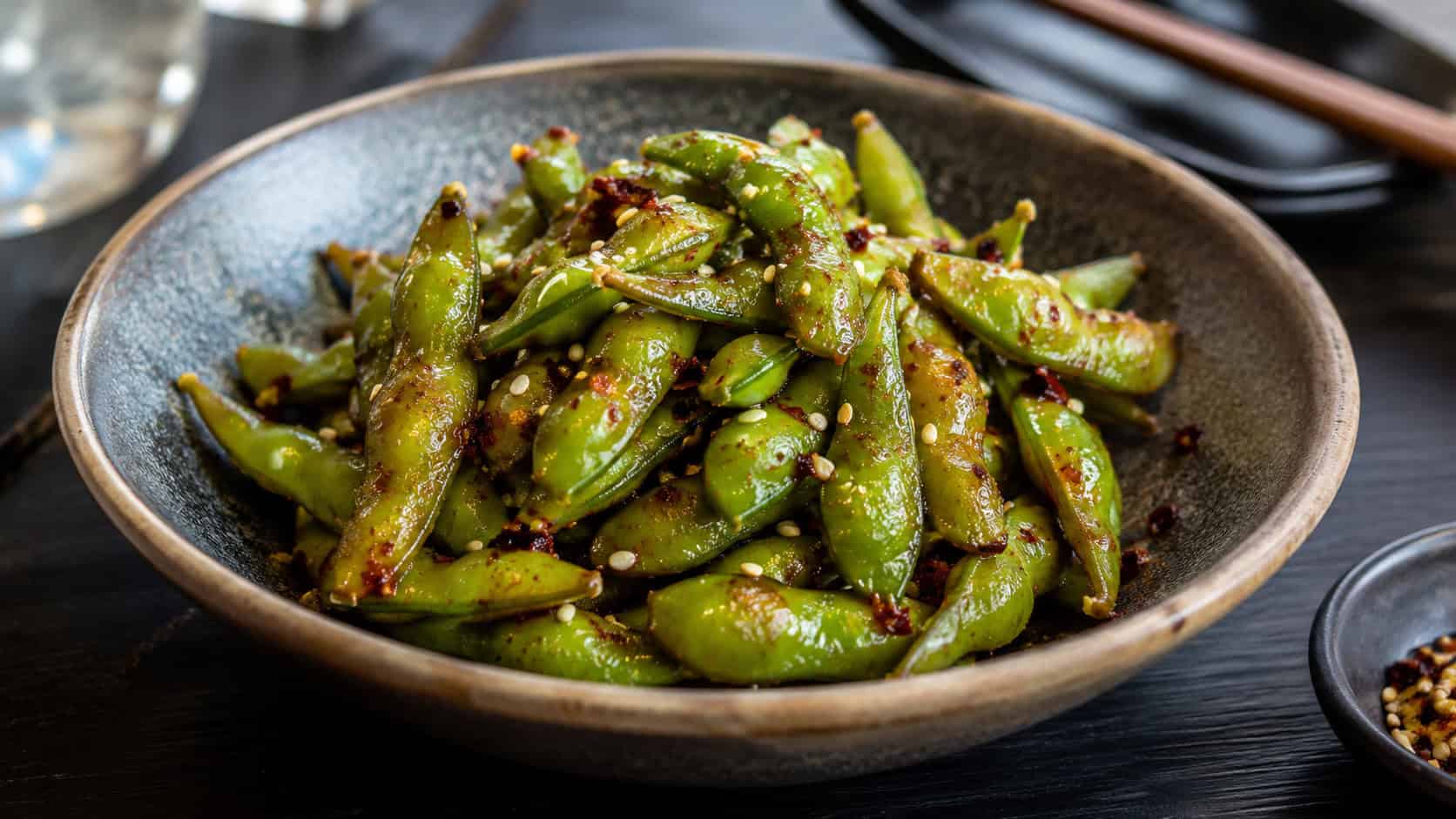 a bowl of glossy spicy edamame pods sprinkled with sesame seeds and chili flakes, in a wide horizontal shot on a dark table, natural light, shallow depth of field, landscape orientation