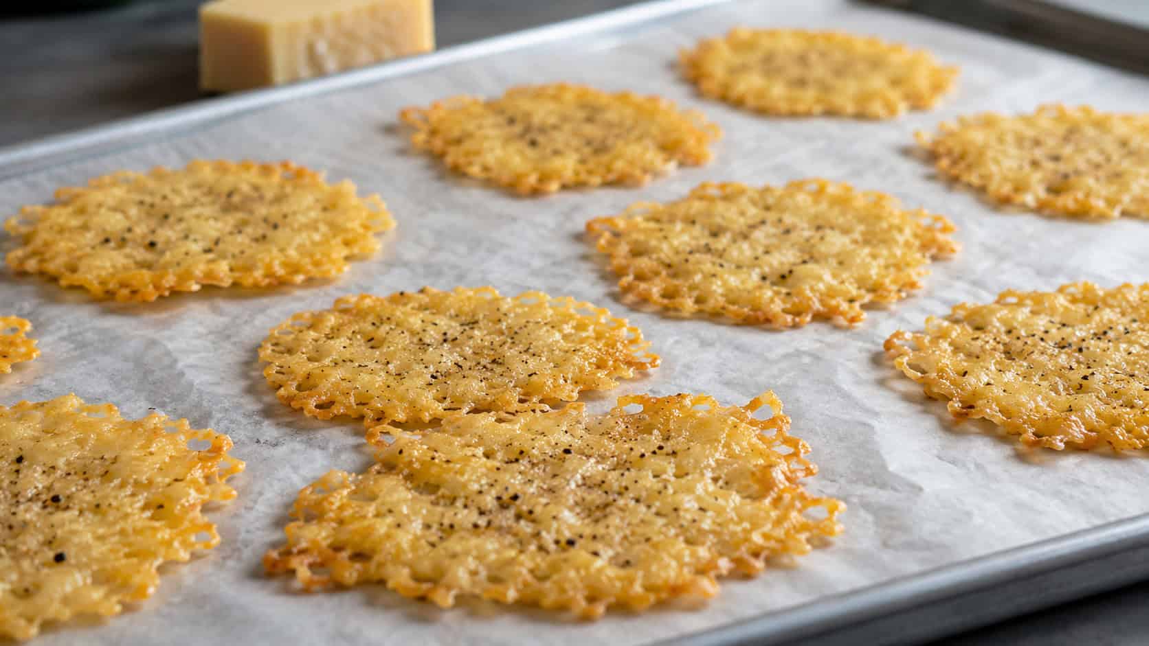 golden lacy parmesan crisps arranged wide in landscape orientation on a parchment-lined tray, crisp and flecked with black pepper, natural light, shallow depth of field, a small wedge of parmesan blurred in the background