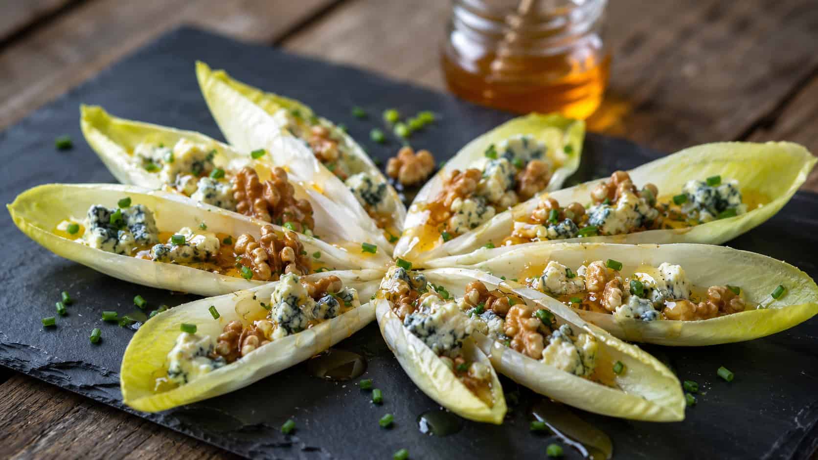endive leaves filled with roquefort and walnuts arranged wide in a landscape fan on a dark slate board, drizzled with honey and sprinkled with chives, natural light, shallow depth of field, a small honey jar blurred behind