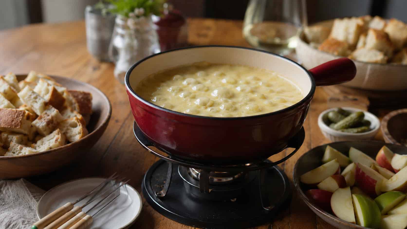 a bubbling pot of swiss cheese fondue shot wide in landscape orientation on a wooden table, surrounded by bowls of crusty bread cubes and apple slices, fondue forks resting nearby, warm natural light, shallow depth of field, soft background blur