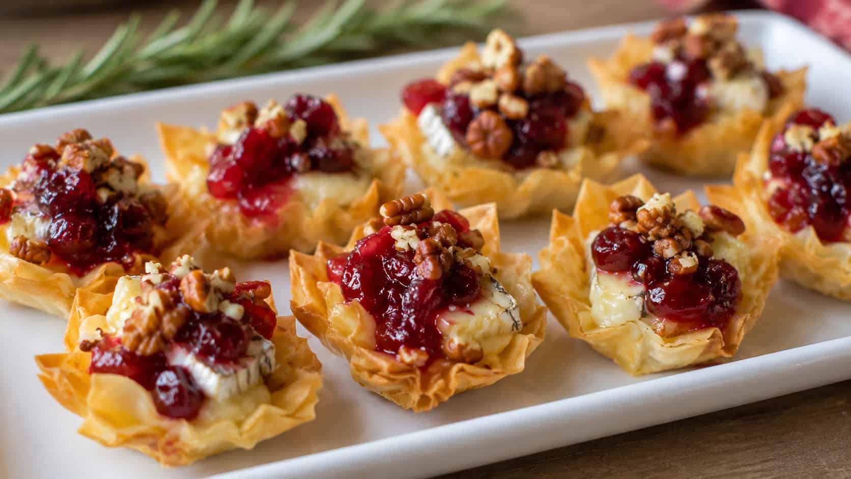 cranberry and brie phyllo bites arranged wide in landscape orientation on a white platter, melty cheese topped with glistening red cranberry and chopped pecans, natural light, shallow depth of field, a sprig of rosemary blurred in the background