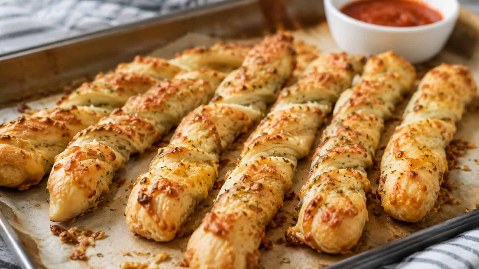 golden asiago breadsticks arranged wide in landscape orientation on a parchment-lined tray, twisted and glossy with melted cheese and herbs, natural light, shallow depth of field, a small bowl of marinara blurred in the background