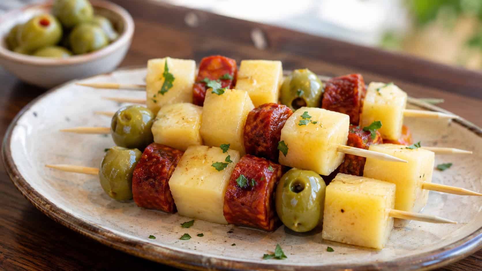 manchego and chorizo skewers arranged wide in landscape orientation on a rustic ceramic plate, golden cheese cubes with red chorizo and green olives, natural light, shallow depth of field, a small dish of olives blurred in the background