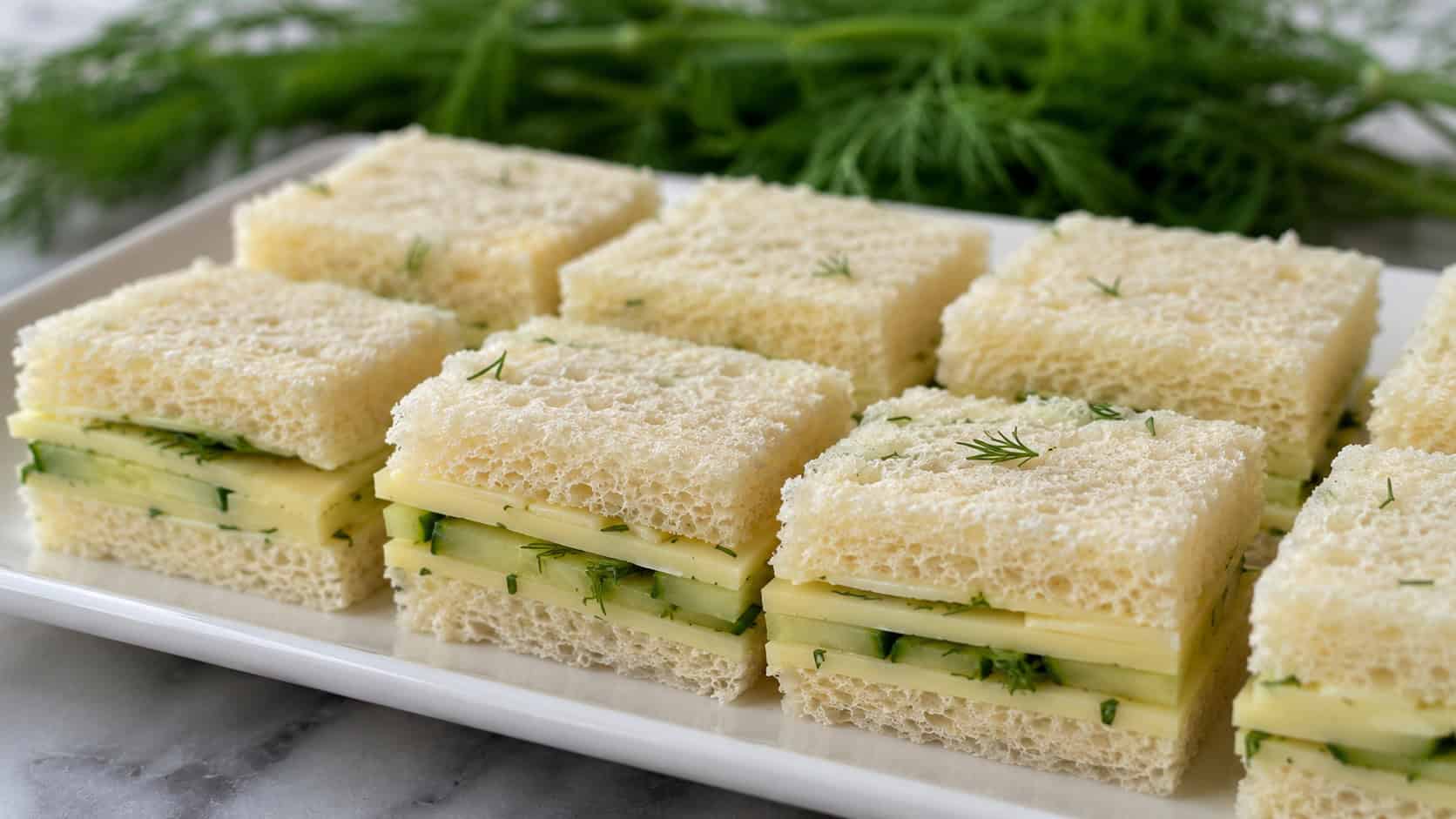havarti and dill cucumber tea sandwiches cut into neat quarters arranged wide in landscape orientation on a white platter, soft layers of cheese and crisp cucumber visible, natural light, shallow depth of field, fresh dill sprigs blurred in the background