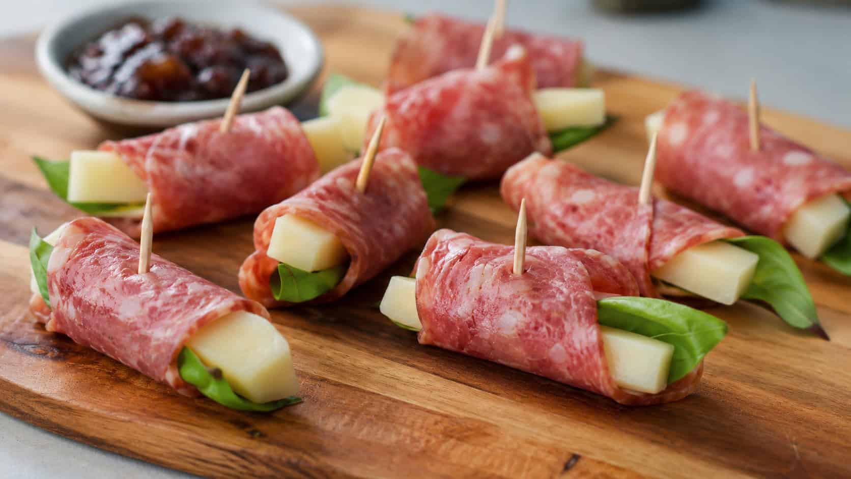 pecorino and salami rolls arranged wide in landscape orientation on a wooden board, rosy salami wrapped around cheese with peeks of basil, secured with toothpicks, natural light, shallow depth of field, a small dish of fig jam blurred behind