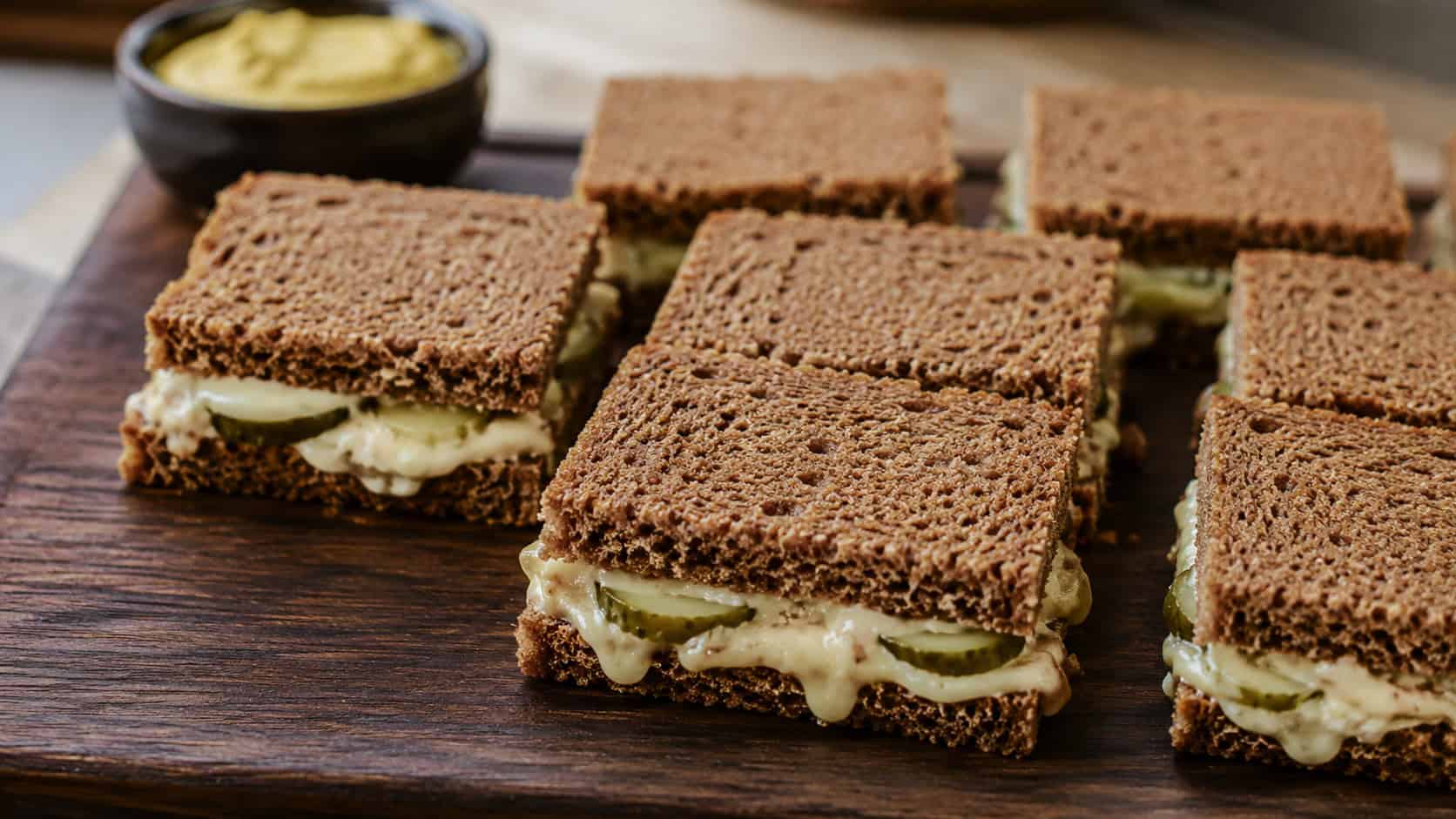 limburger and pickle rye sandwiches cut into neat squares arranged wide in landscape orientation on a dark wooden board, hearty rye with peeks of cheese and pickle, natural light, shallow depth of field, a small dish of mustard blurred behind