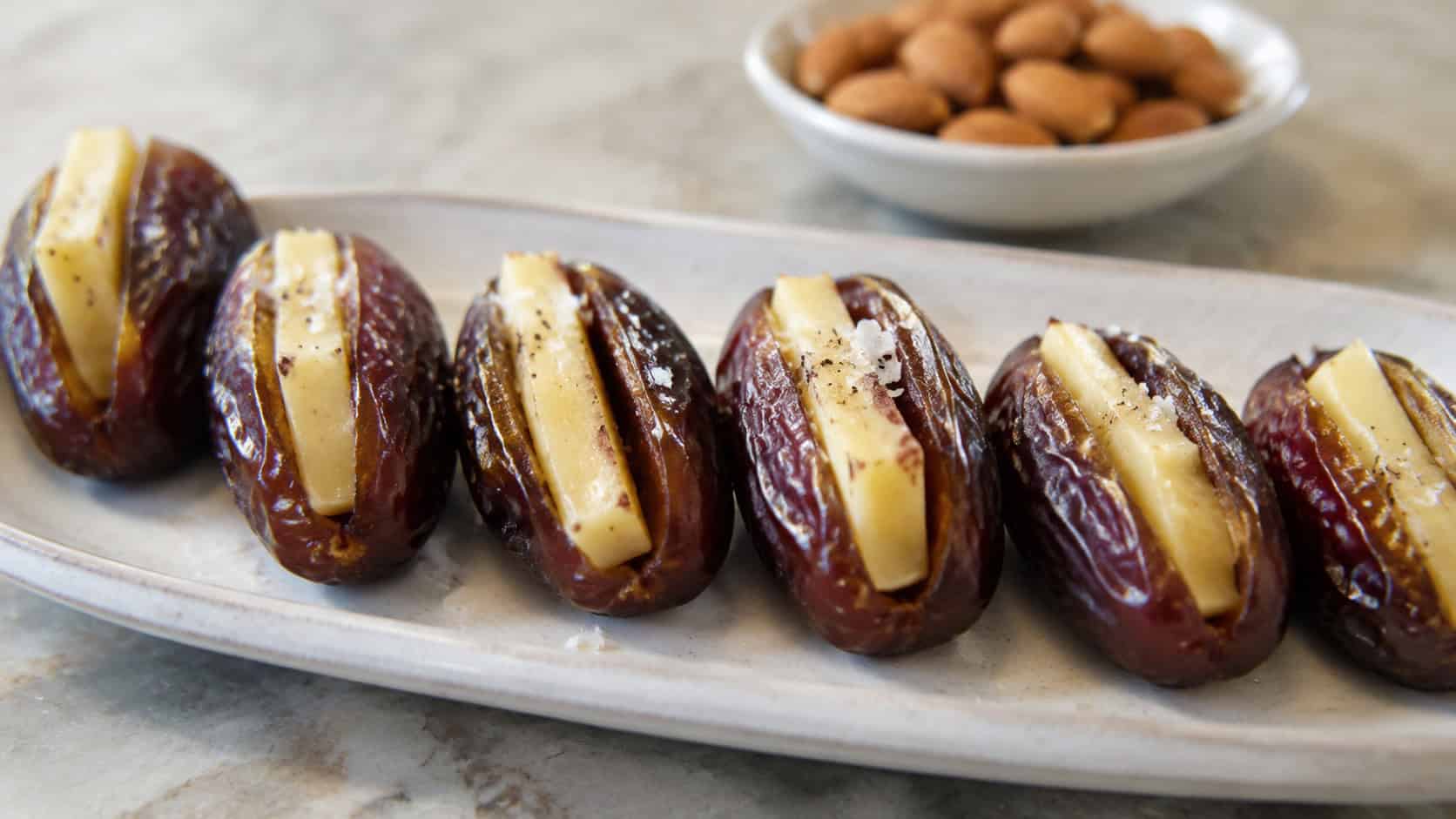 gouda-stuffed Medjool dates arranged in a wide landscape row on a pale ceramic plate, glossy and sprinkled with flaky salt, natural light, shallow depth of field, a small dish of toasted almonds softly blurred behind