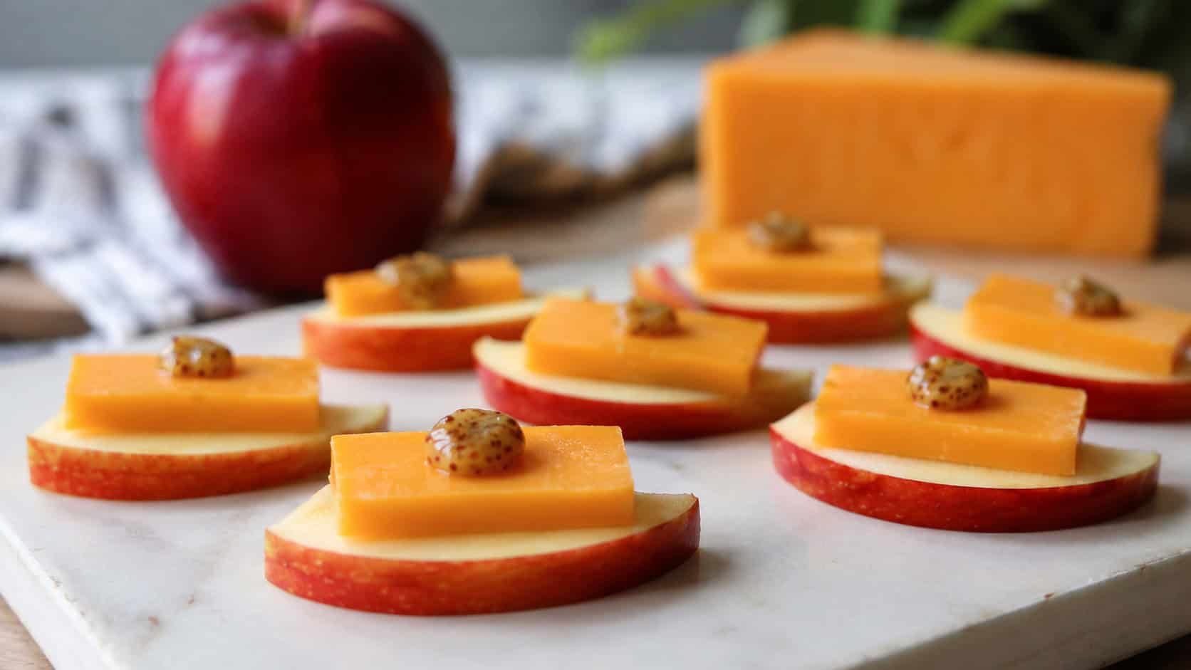 fresh apple slices topped with sharp cheddar arranged wide in landscape orientation on a light cutting board, crisp and colorful with little dots of honey mustard, natural light, shallow depth of field, a whole apple and cheese wedge blurred behind