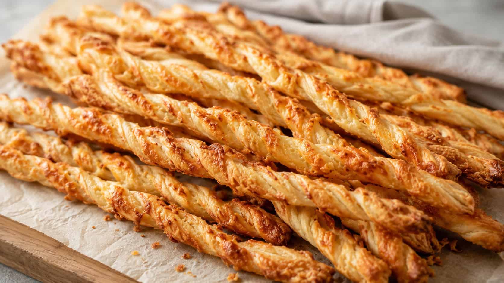 golden twisted gruyère cheese straws piled wide in landscape orientation on a parchment-lined board, flaky and toasted with bits of melted cheese, natural light, shallow depth of field, a linen napkin softly blurred in the background