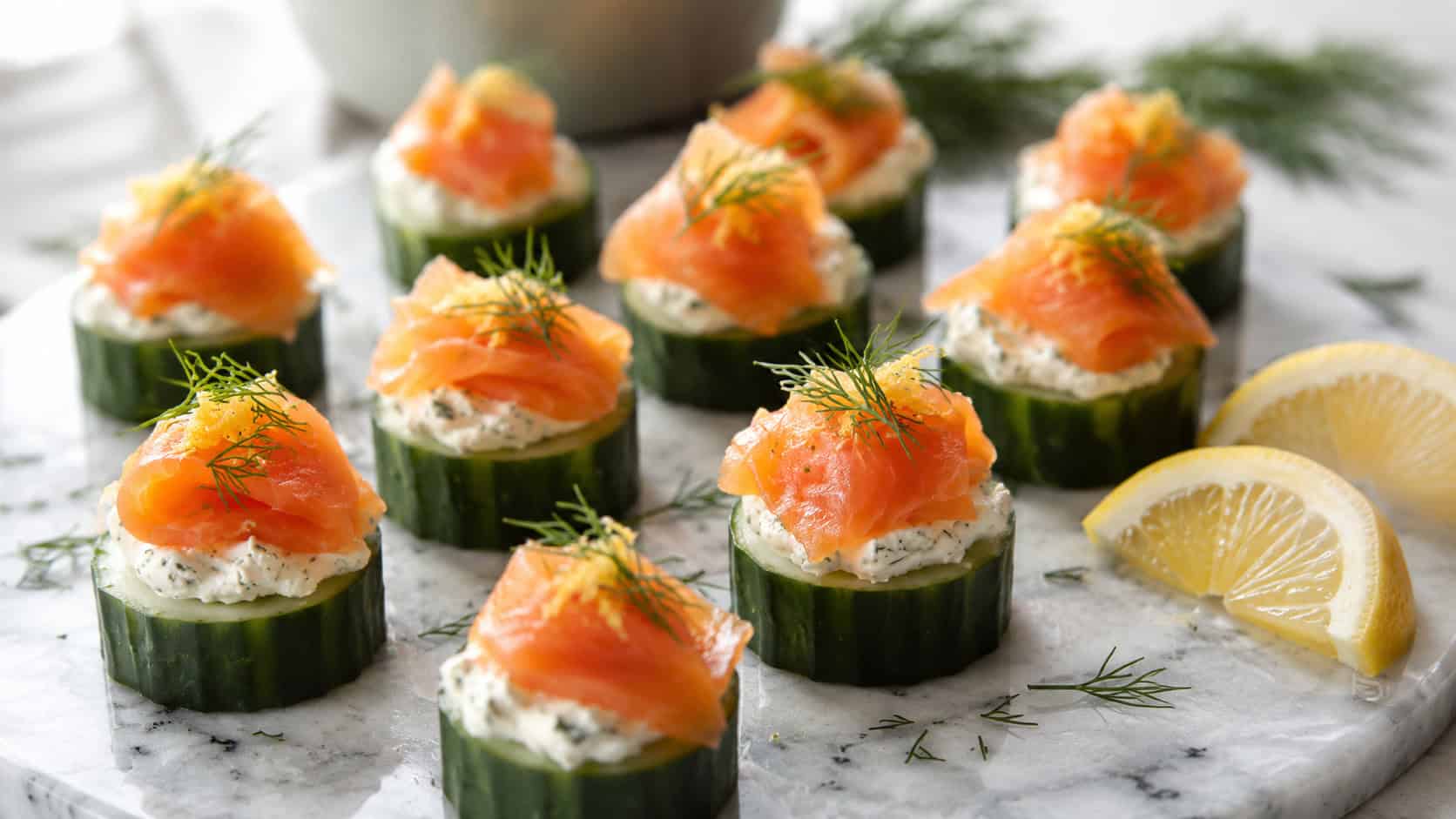 cucumber smoked salmon bites with herbed cream cheese and dill in a wide horizontal shot, arranged on a marble platter with lemon wedges, bright fresh natural light, shallow depth of field, landscape orientation
