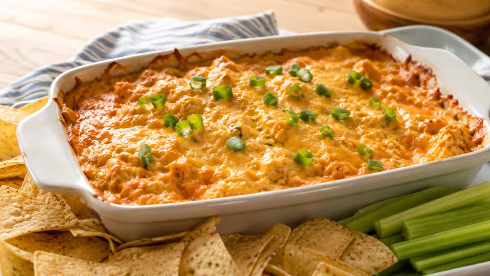 bubbling buffalo chicken dip with melted cheddar and green onion in a wide horizontal shot, served in a baking dish surrounded by tortilla chips and celery, warm natural light, shallow depth of field, landscape orientation