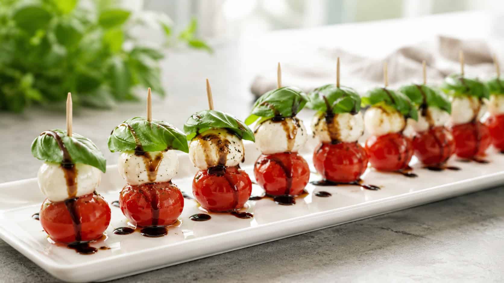 mini caprese skewers with red tomatoes, white mozzarella, and green basil in a wide horizontal shot, lined up on a white platter drizzled with dark balsamic, bright airy natural light, shallow depth of field, landscape orientation