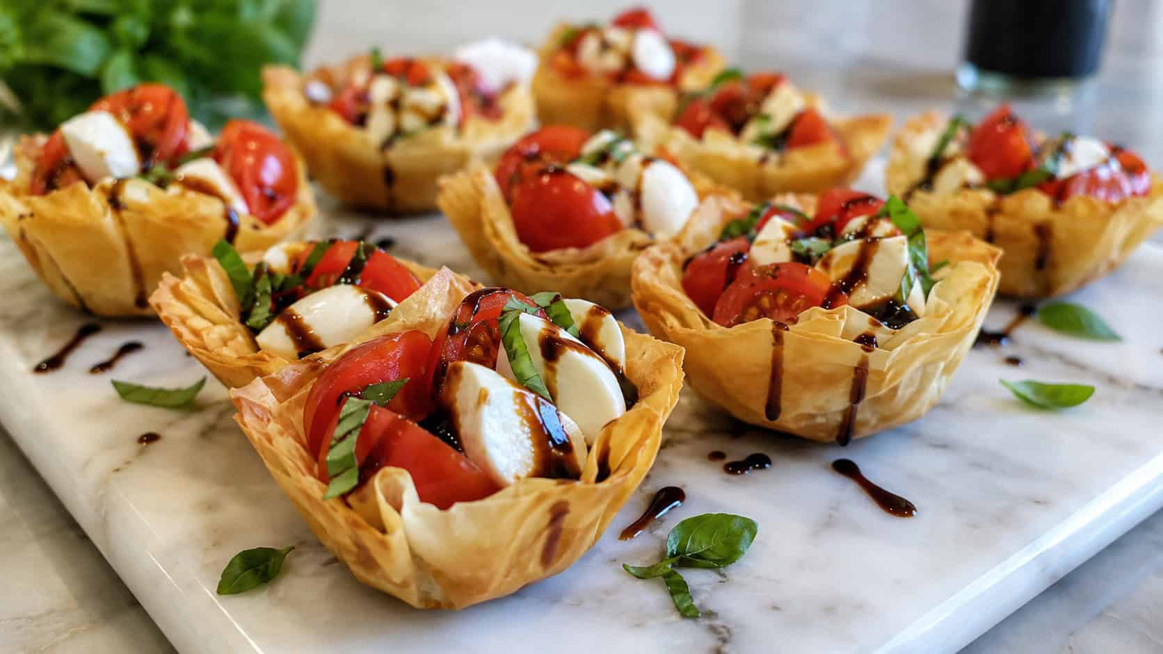 caprese salad cups in crisp phyllo shells filled with tomato, mozzarella, and basil in a wide horizontal shot, drizzled with balsamic on a marble platter, bright natural light, shallow depth of field, landscape orientation