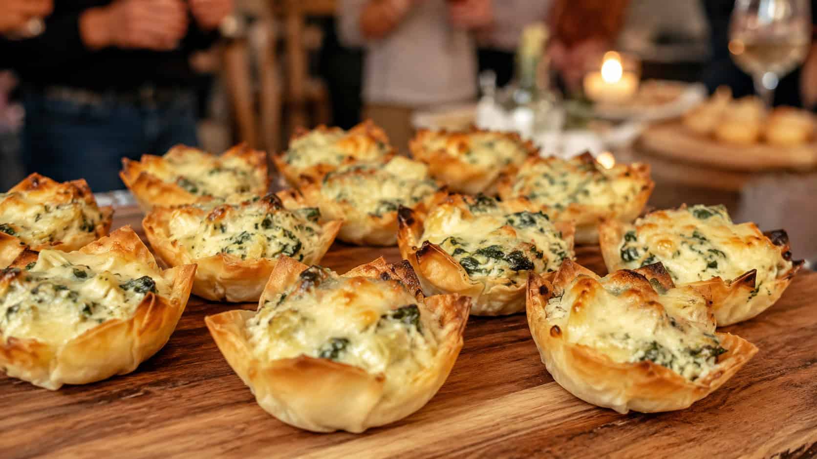 spinach artichoke dip bites in golden phyllo cups with bubbling cheese in a wide horizontal shot, arranged on a wooden board at a party, warm natural light, shallow depth of field, landscape orientation