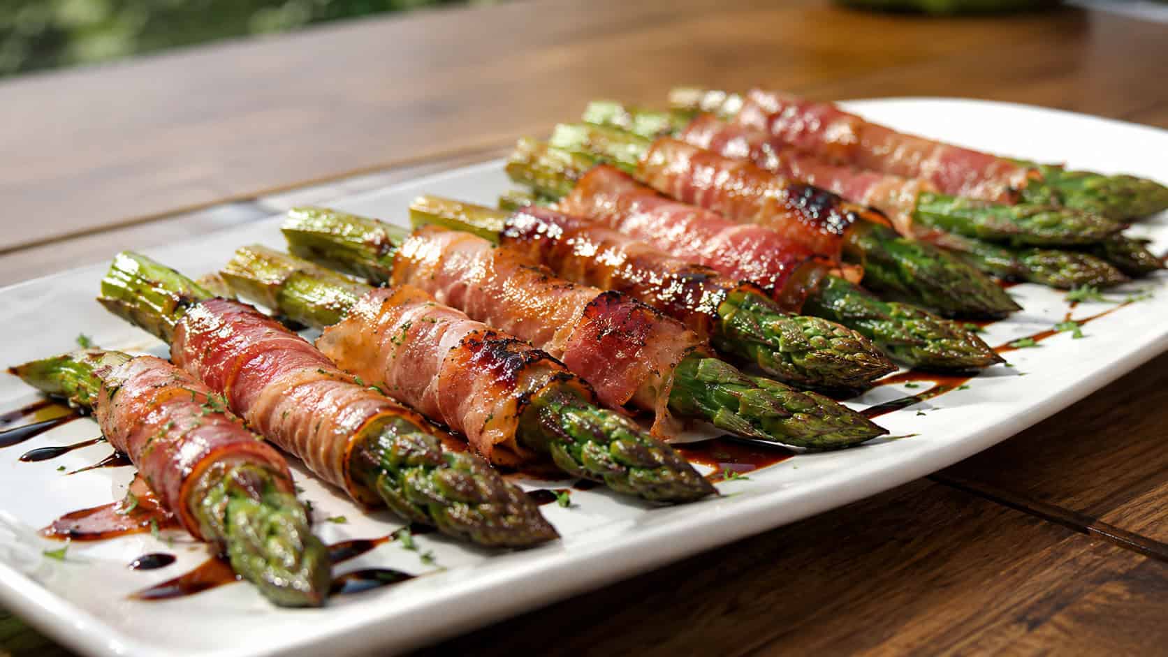 prosciutto-wrapped asparagus spears with crispy edges in a wide horizontal shot, arranged on a long white platter drizzled with balsamic, bright natural light, shallow depth of field, landscape orientation