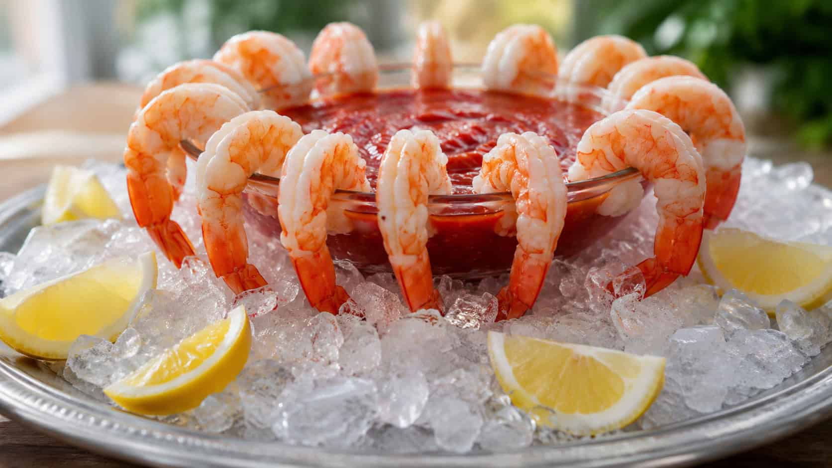 chilled shrimp cocktail hung around the rim of a glass bowl of red sauce in a wide horizontal shot, lemon wedges and crushed ice on a silver platter, bright natural light, shallow depth of field, landscape orientation