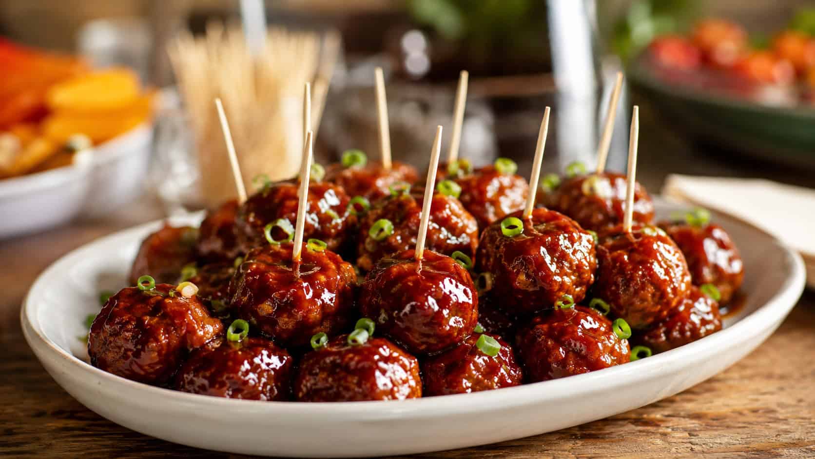 glossy honey BBQ meatballs glistening with sticky sauce in a wide horizontal shot, served in a shallow dish with toothpicks and green onion on a party table, warm natural light, shallow depth of field, landscape orientation