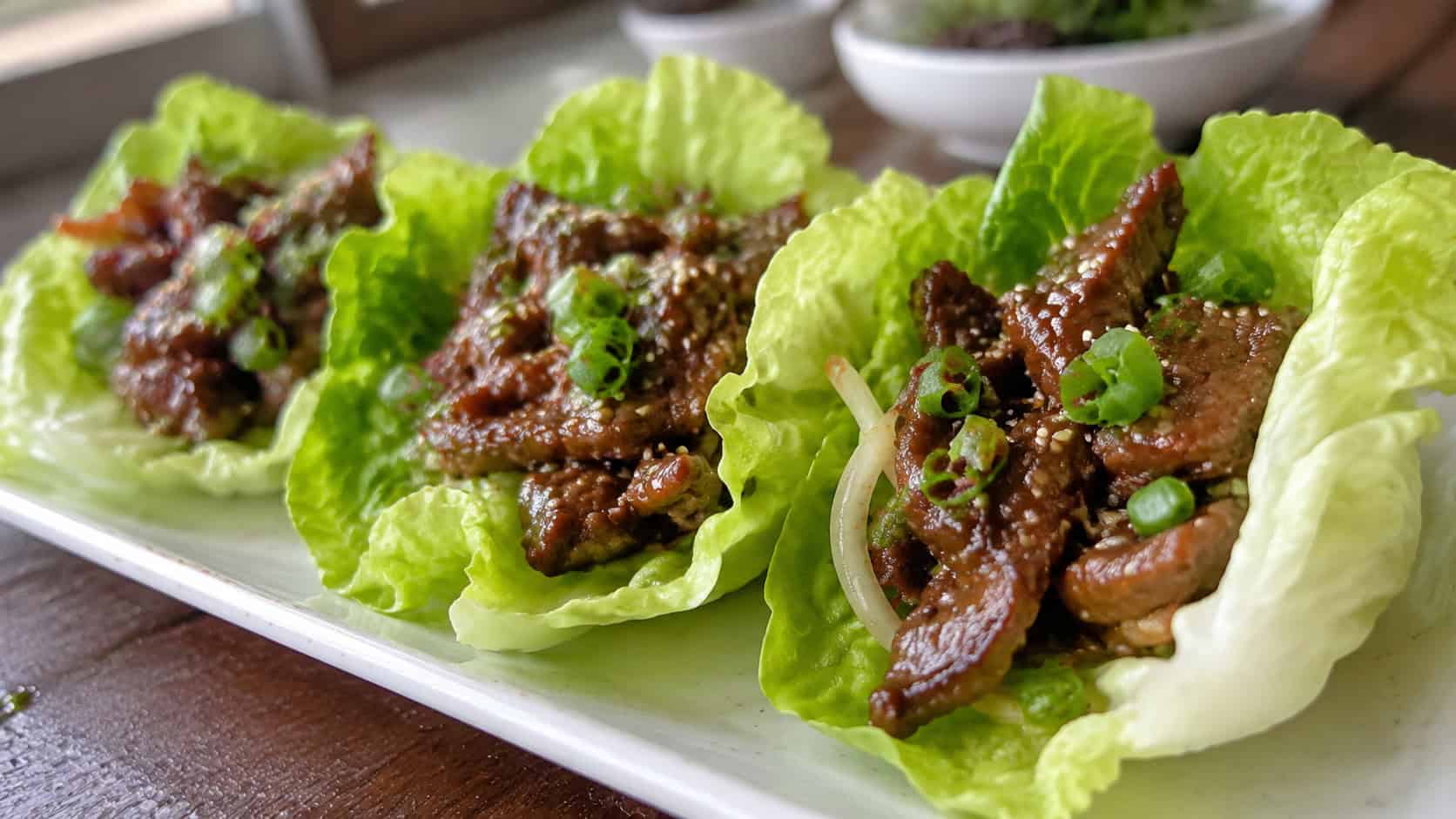 Korean beef lettuce wraps in a wide horizontal shot, crisp green lettuce cups filled with glossy beef, green onions and sesame seeds, on a light platter, natural light, shallow depth of field, landscape orientation