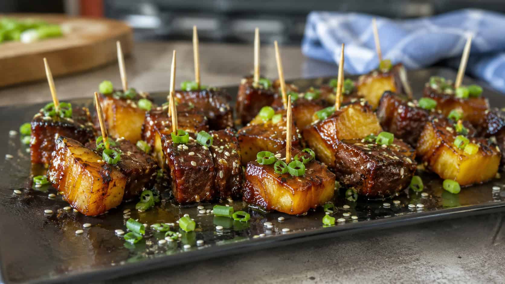 glossy teriyaki beef and pineapple bites on picks in a wide horizontal shot, on a dark platter sprinkled with sesame seeds and green onion, natural light, shallow depth of field, landscape orientation