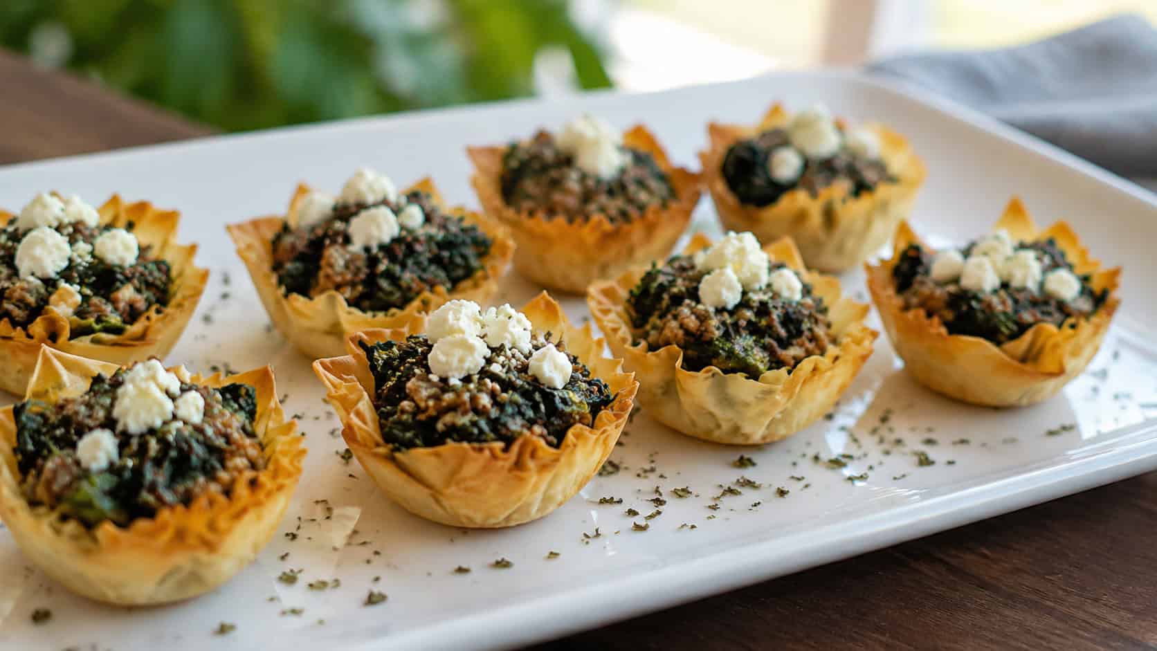 beef and spinach phyllo cups topped with feta in a wide horizontal shot, arranged on a white platter with a sprinkle of oregano, natural light, shallow depth of field, landscape orientation
