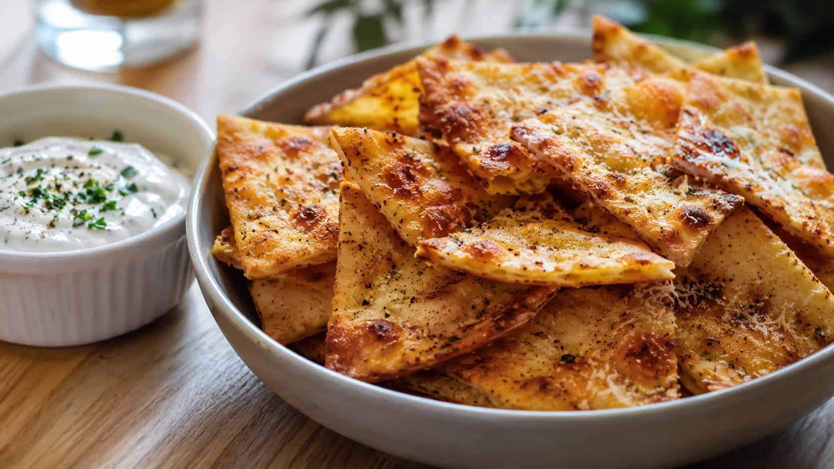 golden garlic parmesan pita chips piled in a shallow bowl in a wide horizontal shot, crisp browned edges and melted cheese flecks, a ramekin of dip beside them, light wooden table, natural light, shallow depth of field, landscape orientation