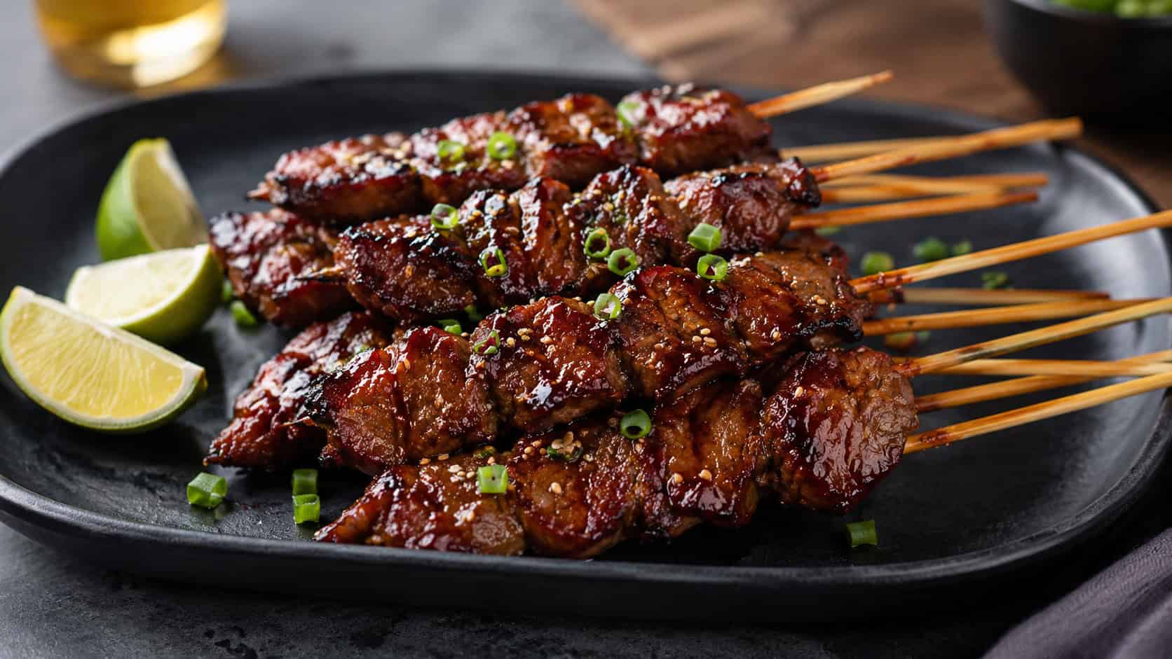 glossy teriyaki beef skewers fanned across a dark platter in a wide horizontal shot, caramelized sticky glaze, sprinkled sesame seeds and green onion, lime wedges nearby, slate surface, warm natural light, shallow depth of field, landscape orientation