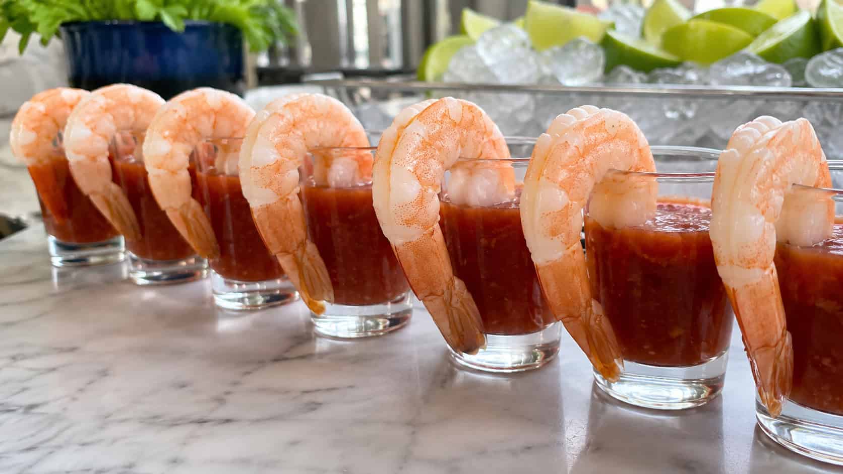 a row of small glasses filled with red cocktail sauce and chilled shrimp hooked over the rims in a wide horizontal shot, lime wedges and ice in the background, marble countertop, bright natural light, shallow depth of field, landscape orientation