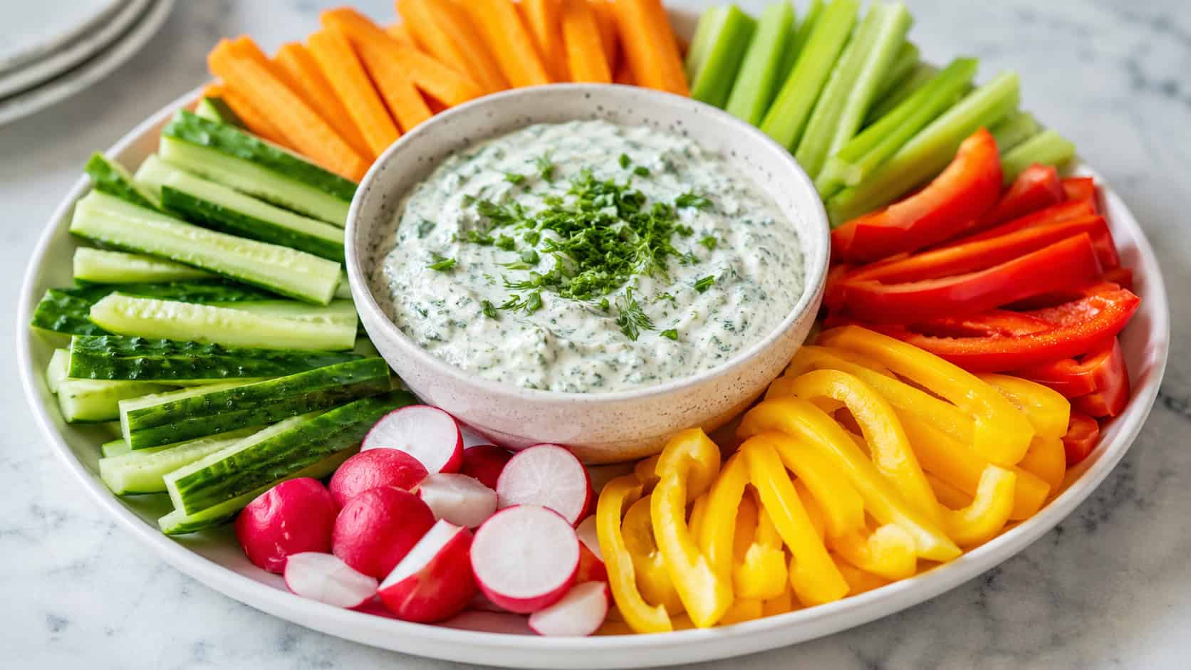a vibrant crudités platter in a wide horizontal shot, colorful carrot, pepper, cucumber and celery sticks fanned around a bowl of creamy green-flecked herb dip, light marble surface, bright natural light, shallow depth of field, landscape orientation