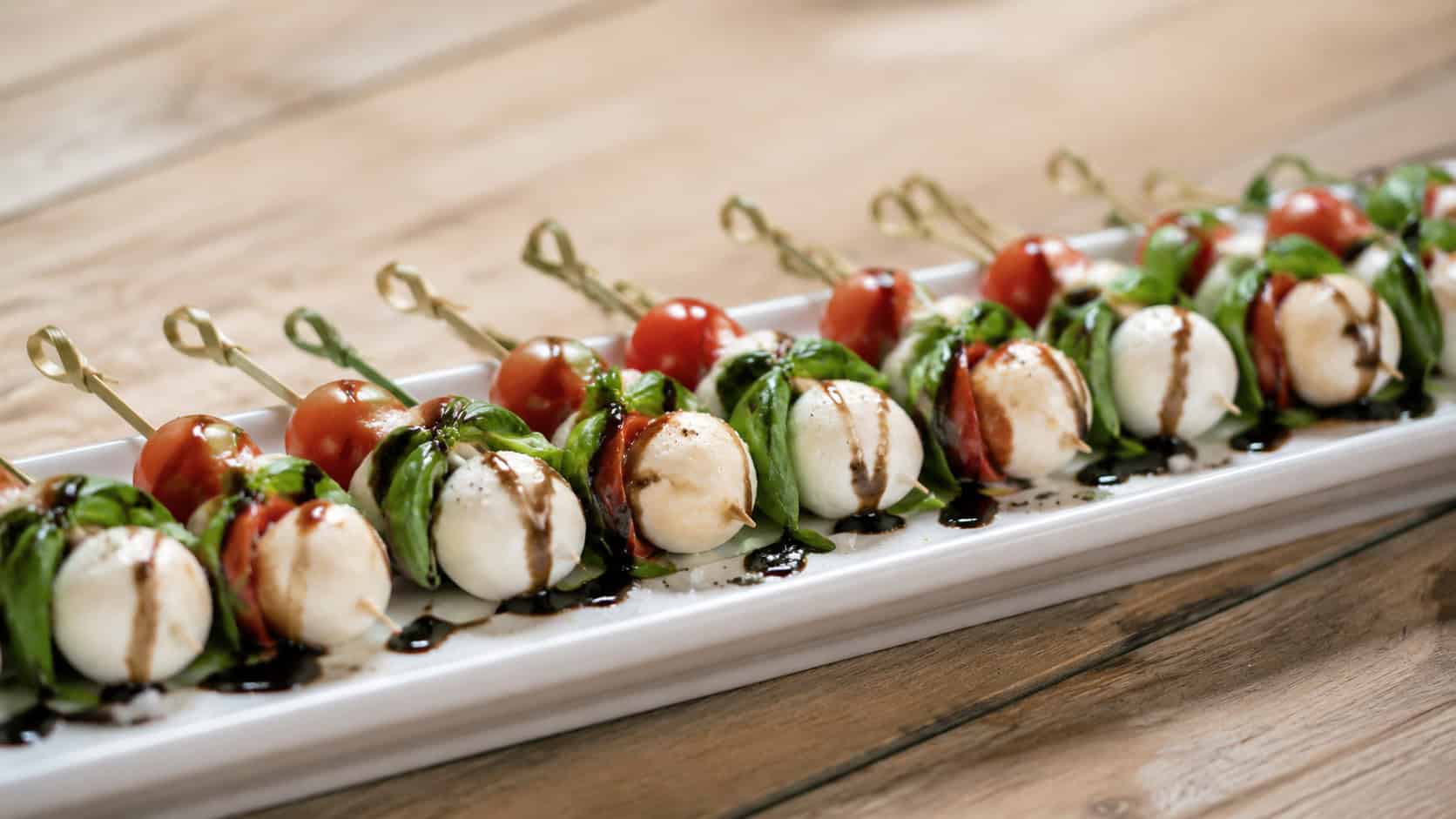 mini caprese skewers lined up on a long white platter in a wide horizontal shot, glossy balsamic drizzle and bright basil, scattered flaky salt, light wooden table, soft natural light, shallow depth of field, landscape orientation